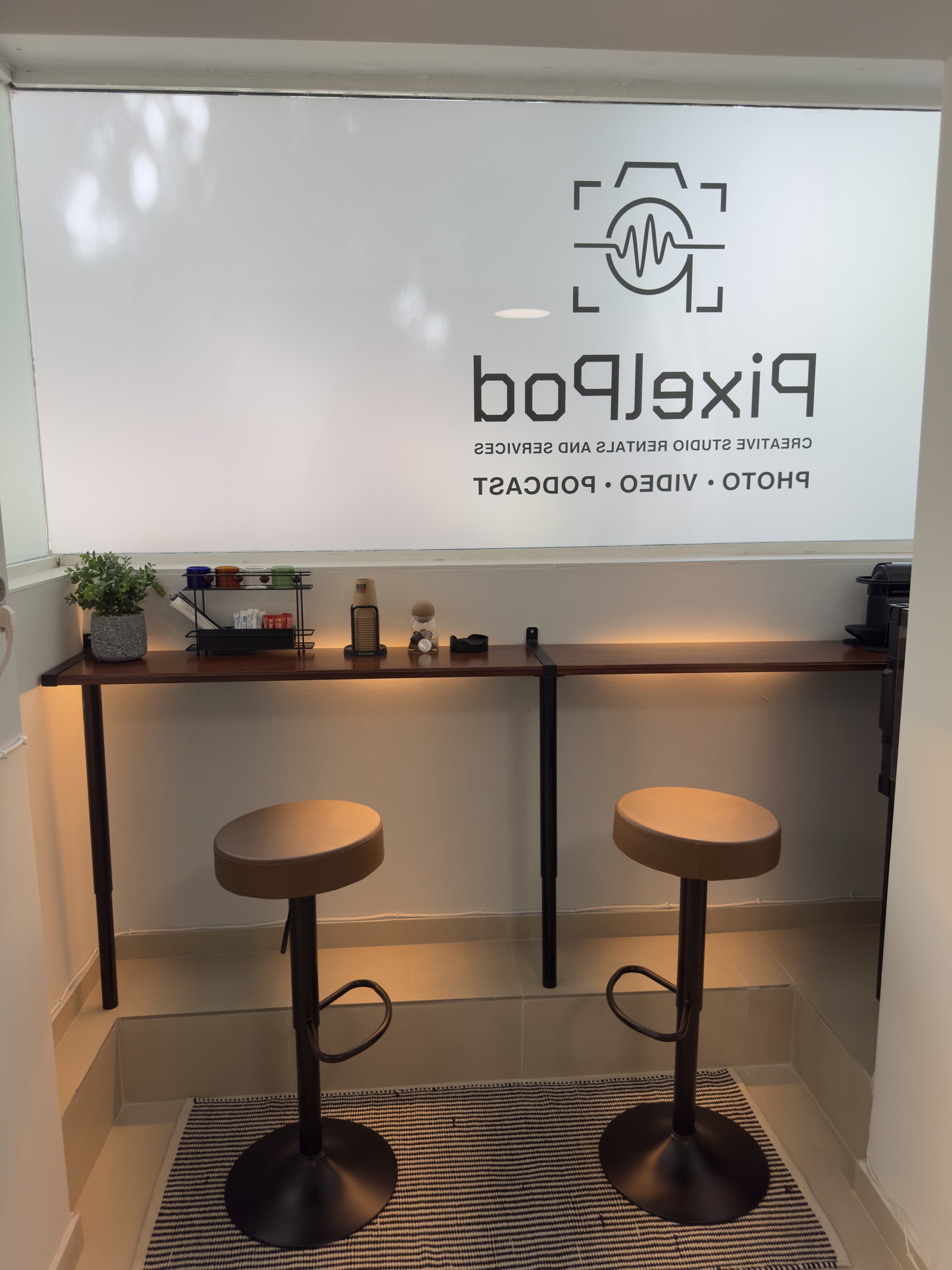 A small breakfast bar area with two high stools, a black table with a plant on the left, a small shelf with condiments, and a window with the logo of Pixeld, a studio specializing in creating studio rentals and services, displayed in reverse. The logo features a camera with a waveform inside.