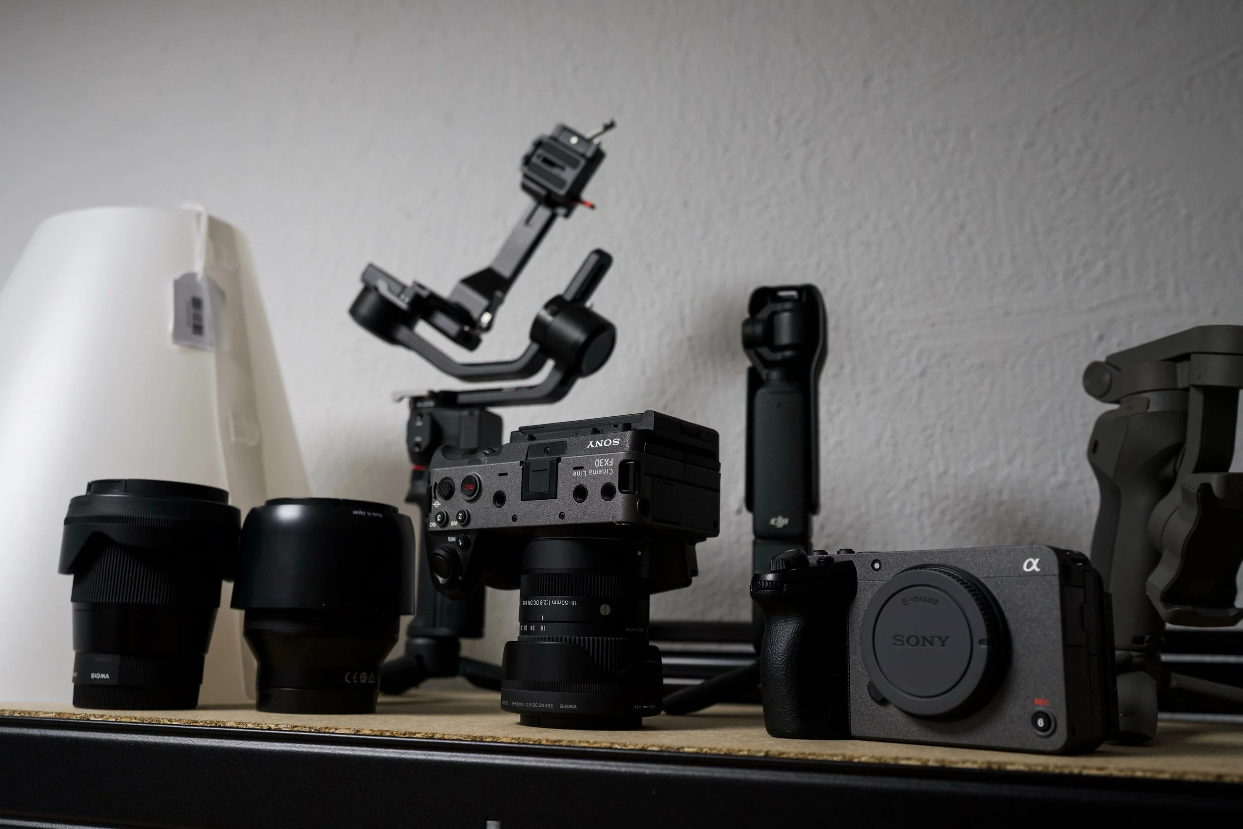 Various camera lenses, a camera gimbal stabilizer, and a Sony mirrorless camera on a shelf against a light-colored wall.