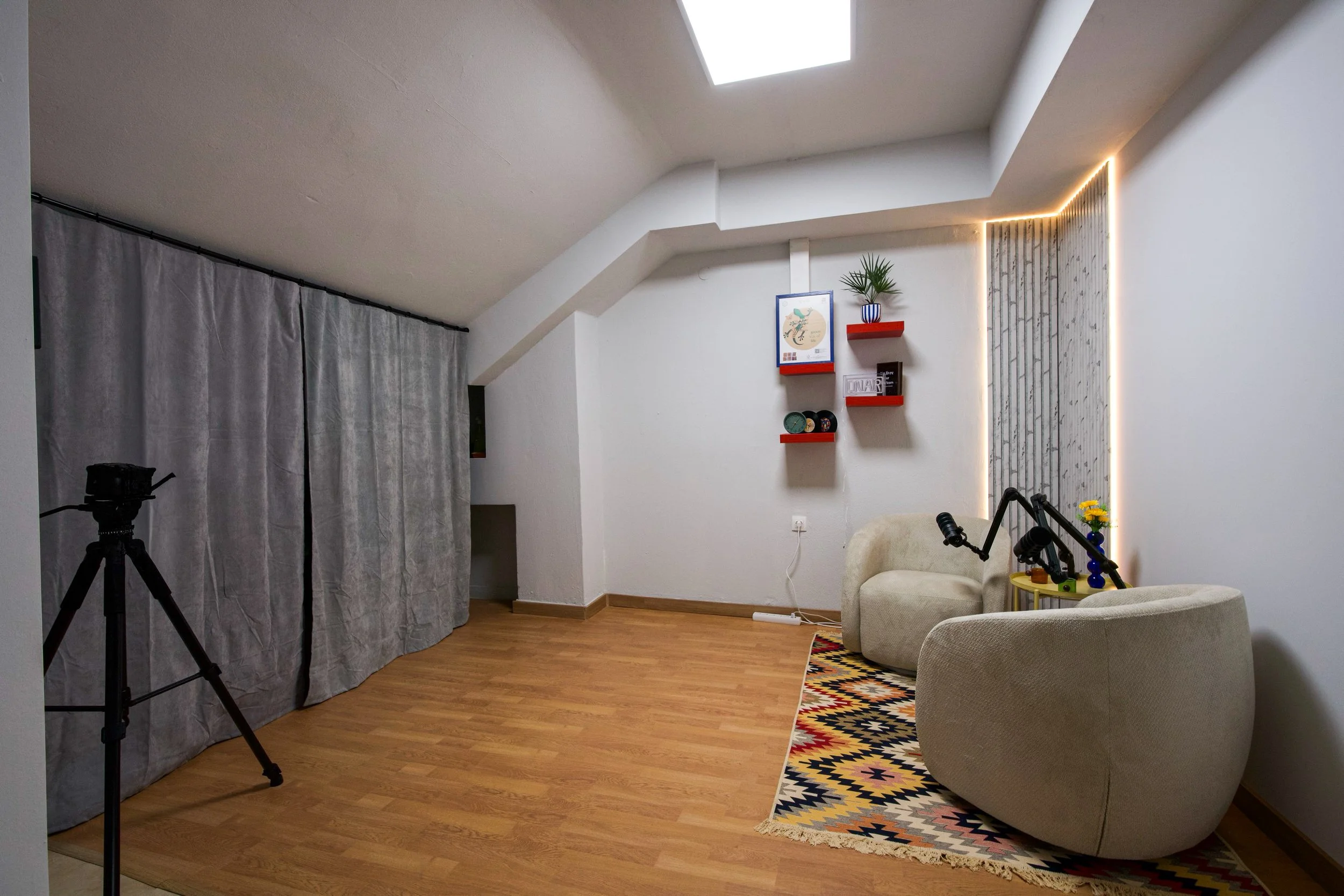 A cozy living room with a skylight, a beige armchair, colorful patterned rug, wall decorations, and camera equipment, including a tripod.