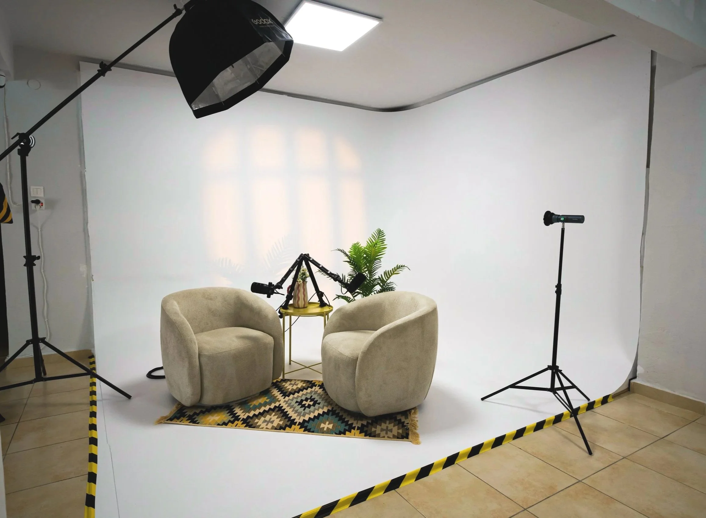 A photography setup with a white backdrop, two beige armchairs facing each other, a small yellow table with a plant, multiple studio lights and equipment, all surrounded by black and yellow caution tape on the floor.
