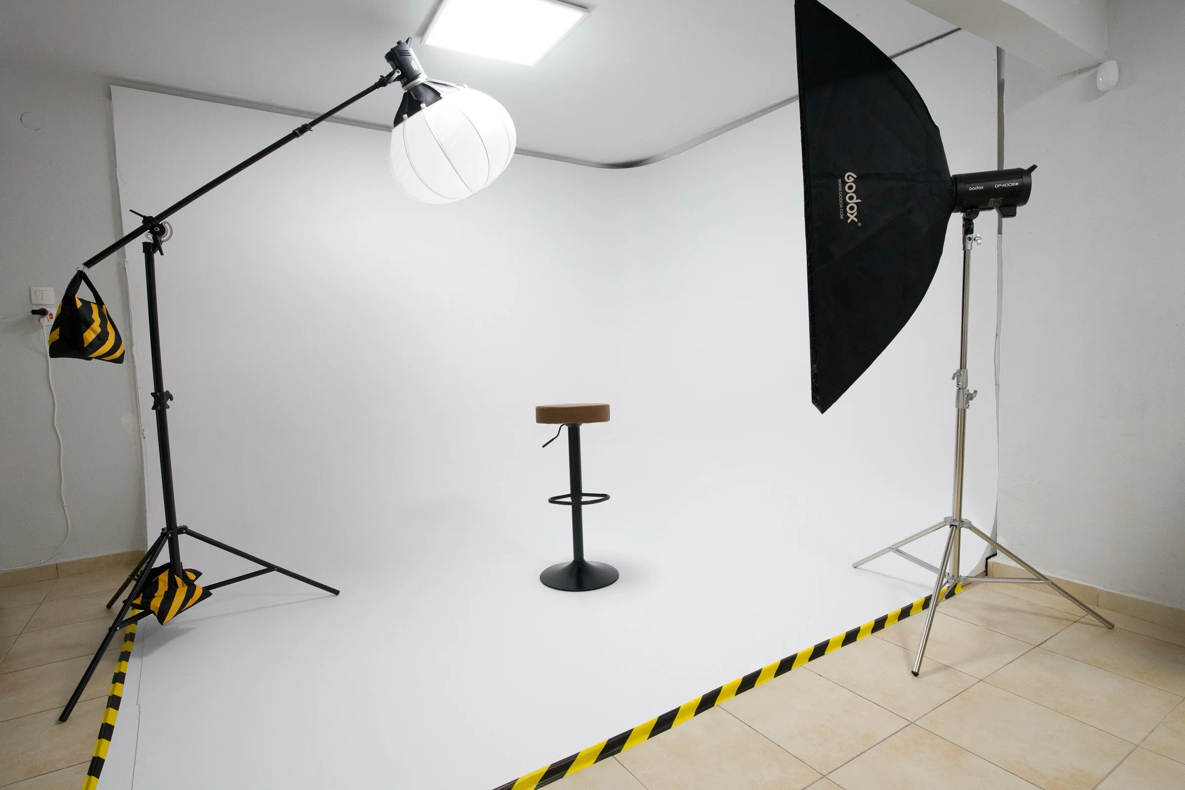 Photography studio setup with a white backdrop, studio lights, light modifiers, and a stool, all surrounded by yellow and black caution tape.