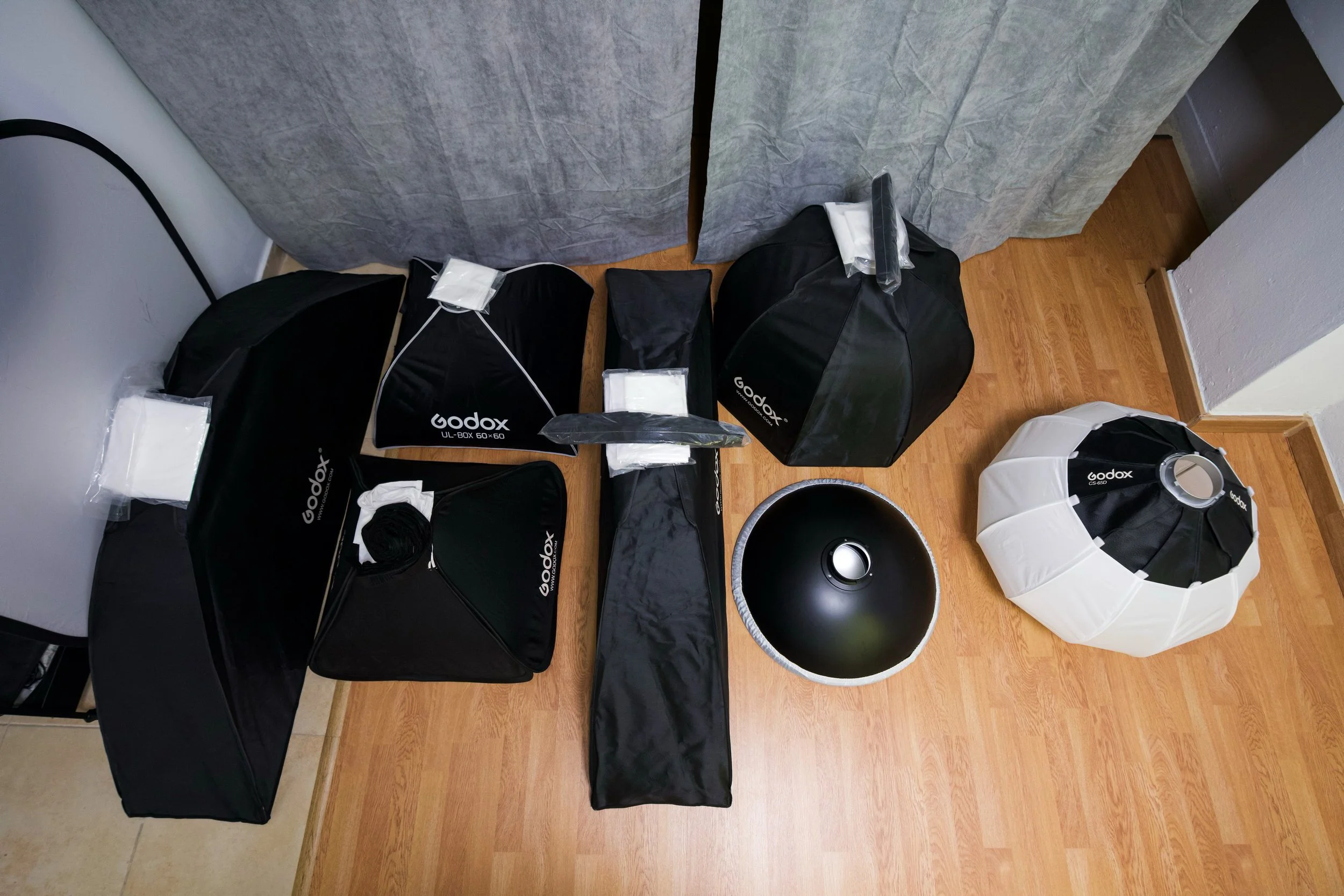 Collection of photography equipment including softboxes, studio lights, and reflectors arranged on a wooden floor and against a wall.