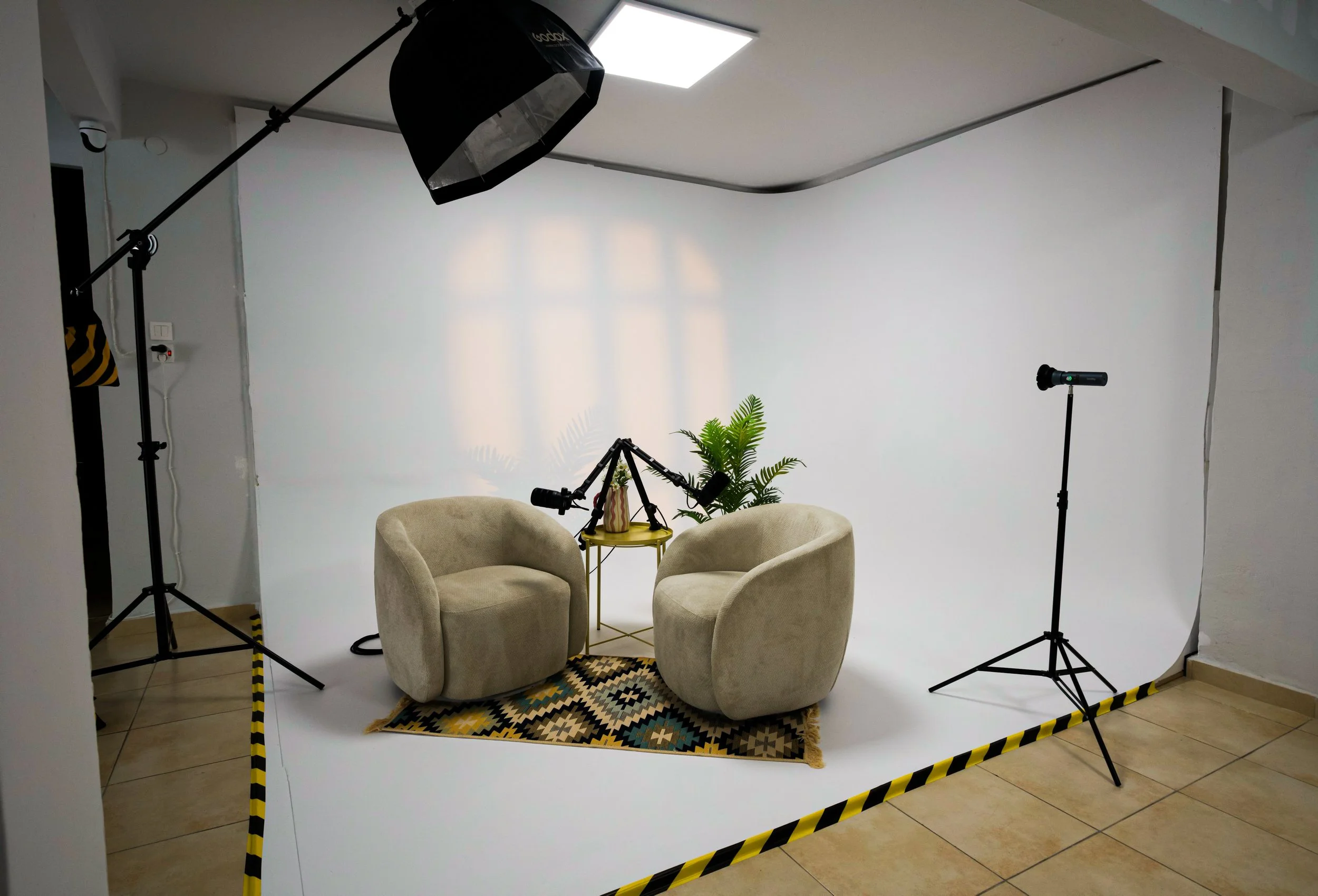 A small photography setup with two beige armchairs facing each other, a small yellow table with a potted plant on it, and a patterned rug underneath. The setup is enclosed by black and yellow striped tape, with studio lights and microphones positioned around the scene, indicating a recording or interview session in a room with white walls and tile flooring.