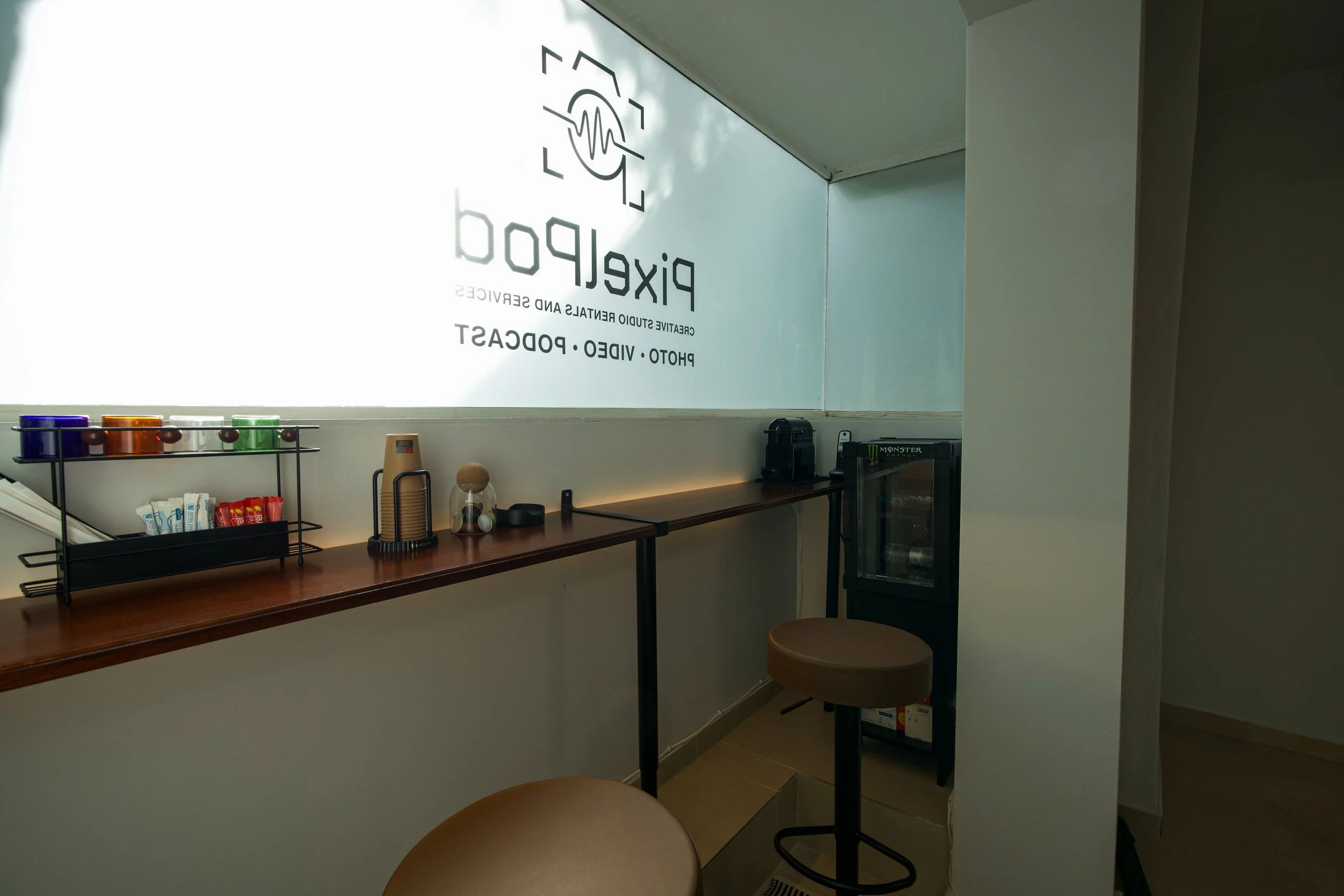 Interior of a cafe or coffee shop with a wooden counter, a black shelf with condiment packets, small appliances, a mini fridge, and a round stool, with a large window and signage that reads 'Pixelpob' and 'Creating Studio Rentals and Services'.