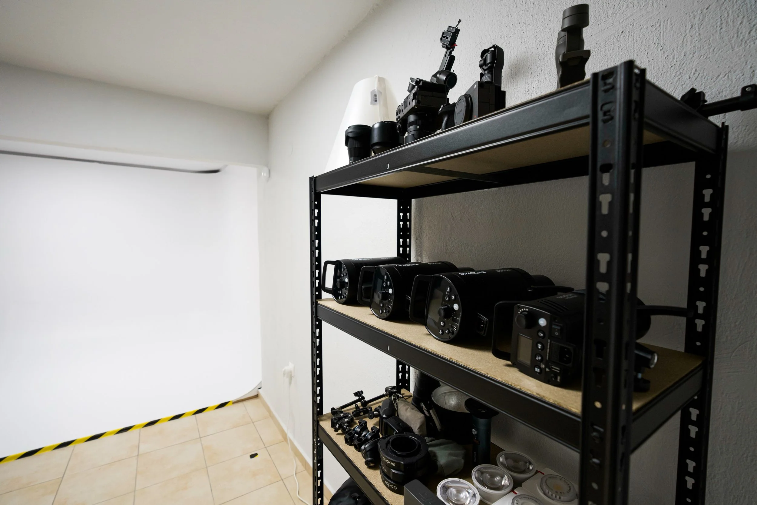 Black metal shelving unit with photography and videography equipment including cameras, lenses, and lighting accessories, against a plain white wall with a tiled floor.