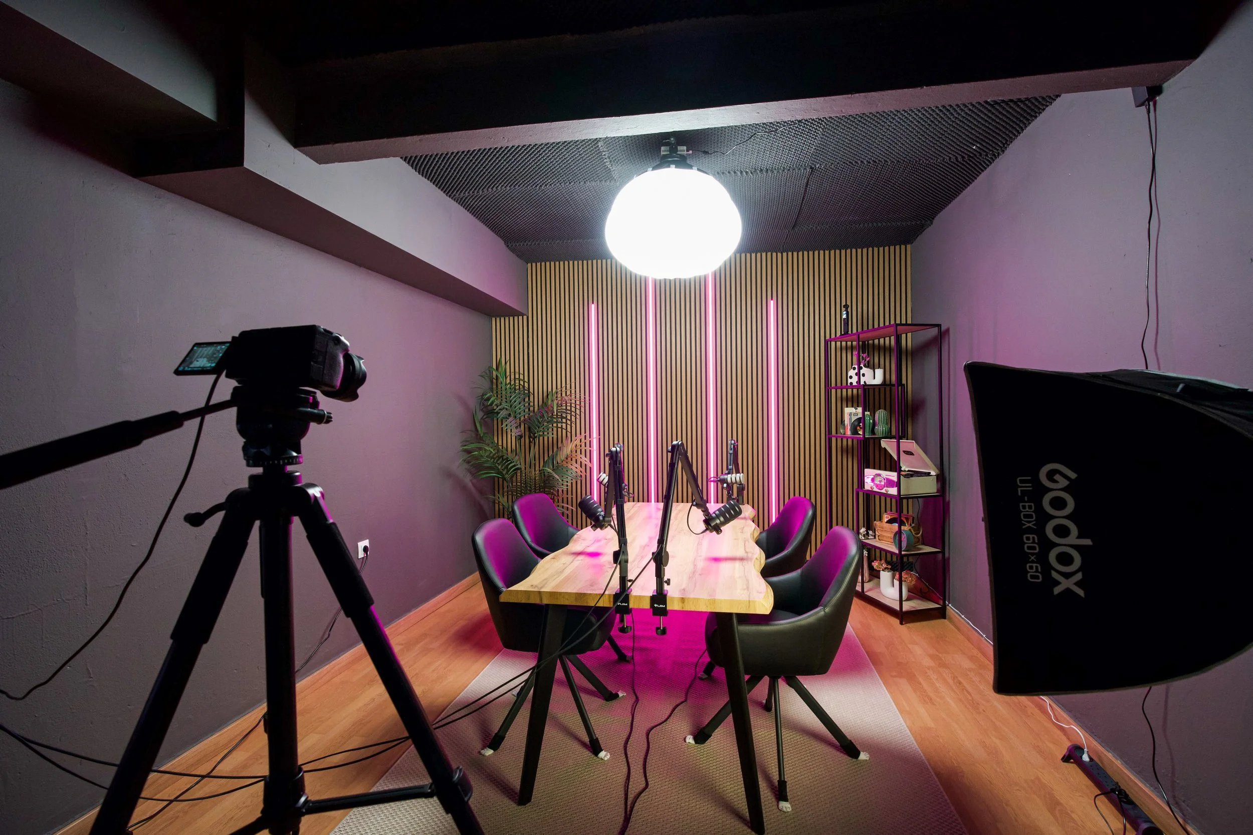 Indoor recording studio with a wooden table, six black chairs, a large bright light fixture hanging from the ceiling, a camera on a tripod, a pink shelf with decorative items, and a wooden slat wall with pink neon lights.