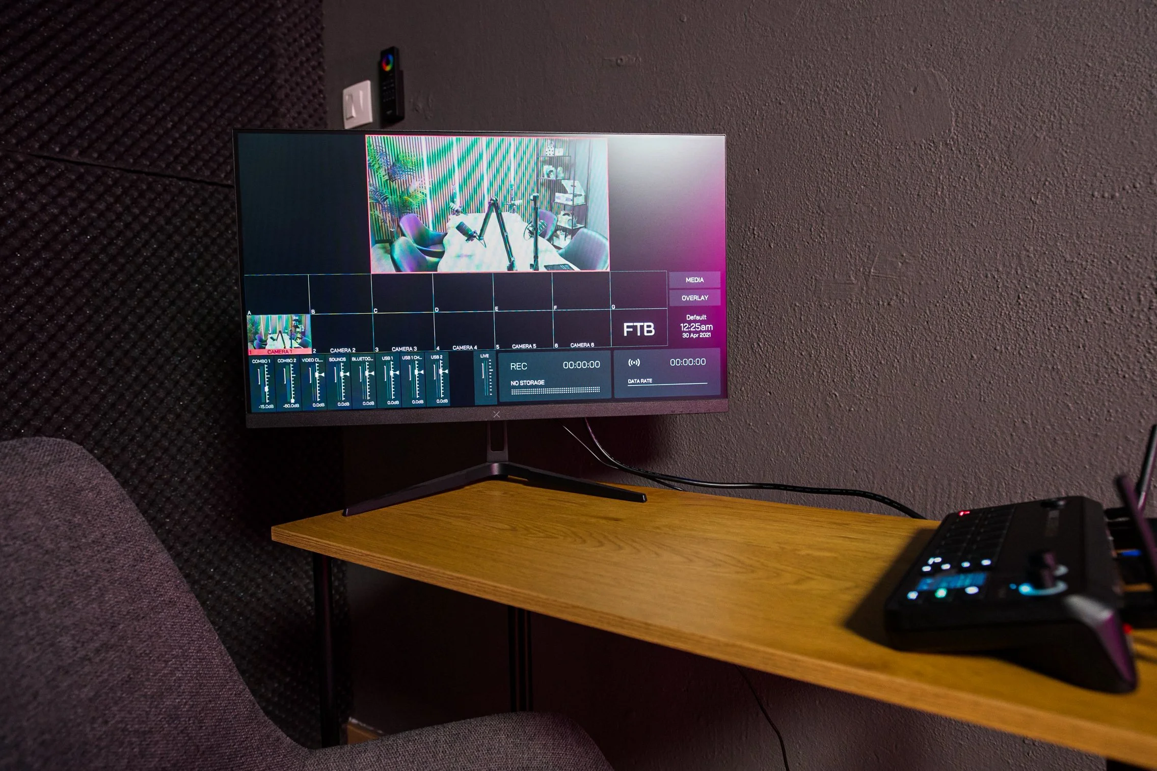 A computer monitor displaying a studio recording setup with microphones and chairs in a room with acoustic panels. The monitor is on a wooden table against a textured wall.