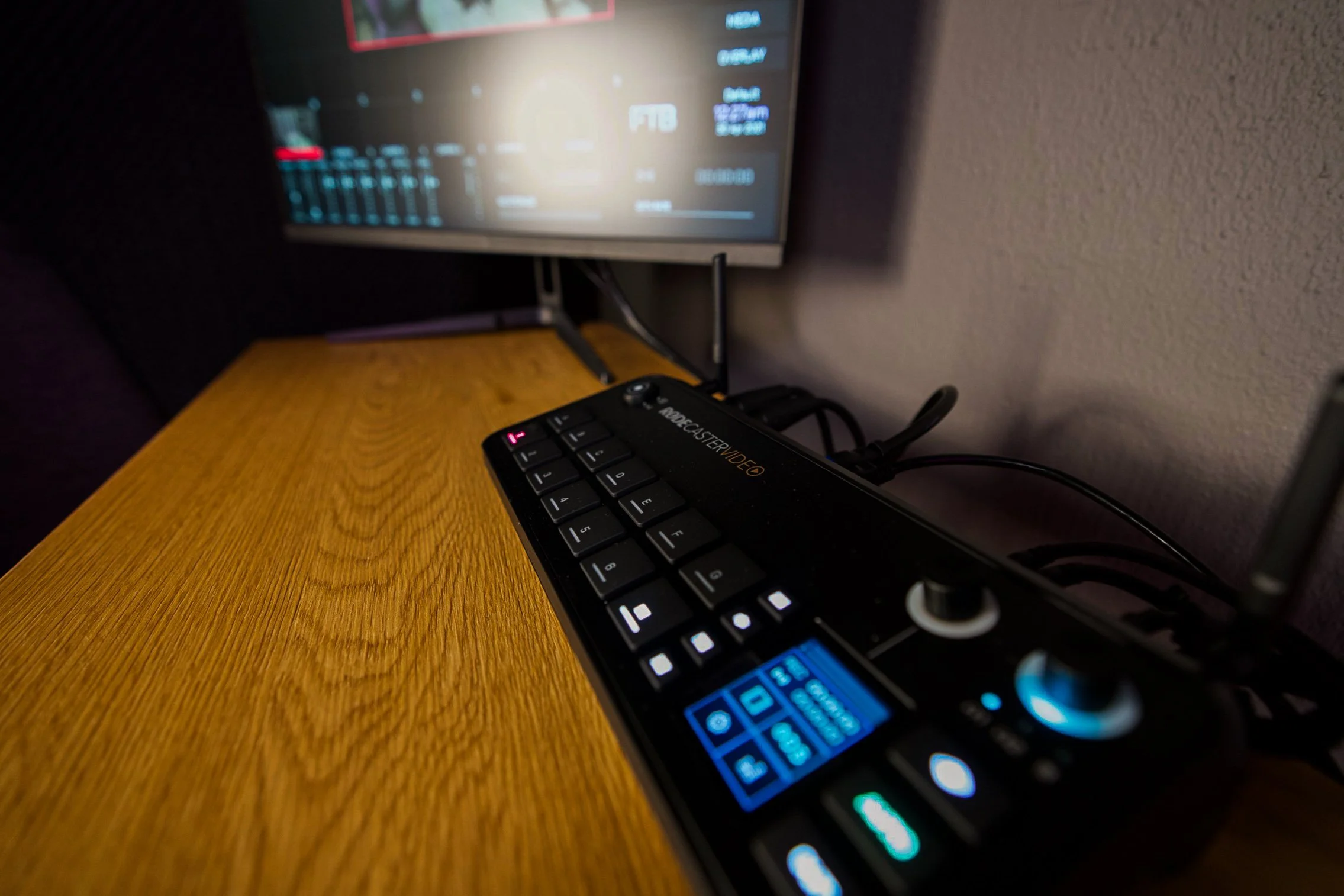 A close-up of a black keyboard connected to an audio/video receiver placed on a wooden surface. In the background, a monitor displays a digital interface with various settings.