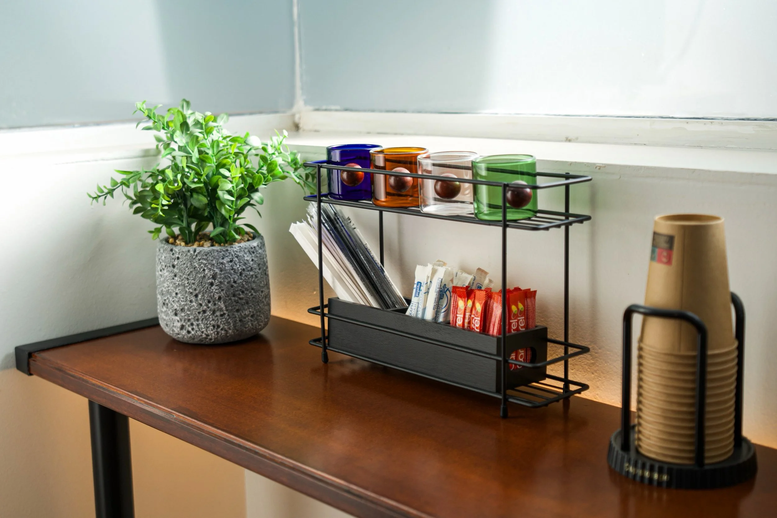A wooden table with a textured gray potted plant, colorful glass jars, a black wire rack with coffee supplies, a roll of paper towels, and a small cup.