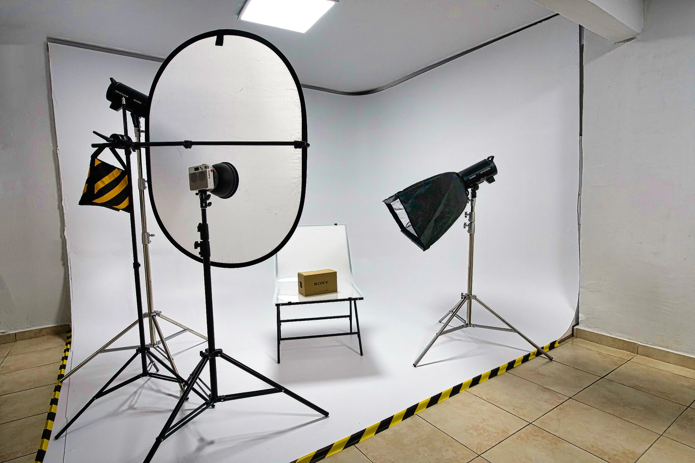 Photography studio setup with white backdrop, studio lights, softboxes, a reflector, and a small table with a Sony product box, surrounded by yellow and black caution tape on the floor.