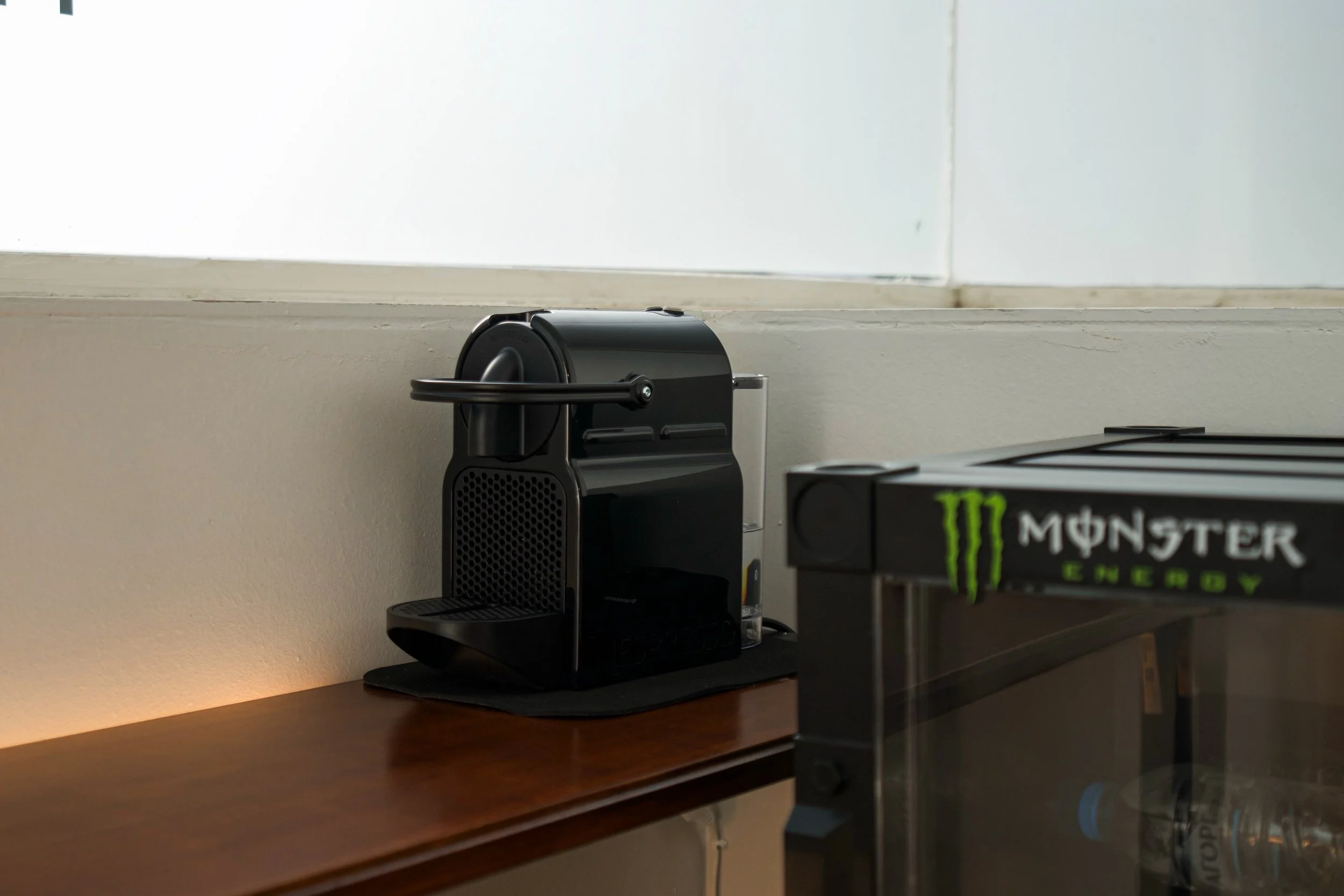 Black coffee maker on a wooden desk next to a Monster Energy drink can.