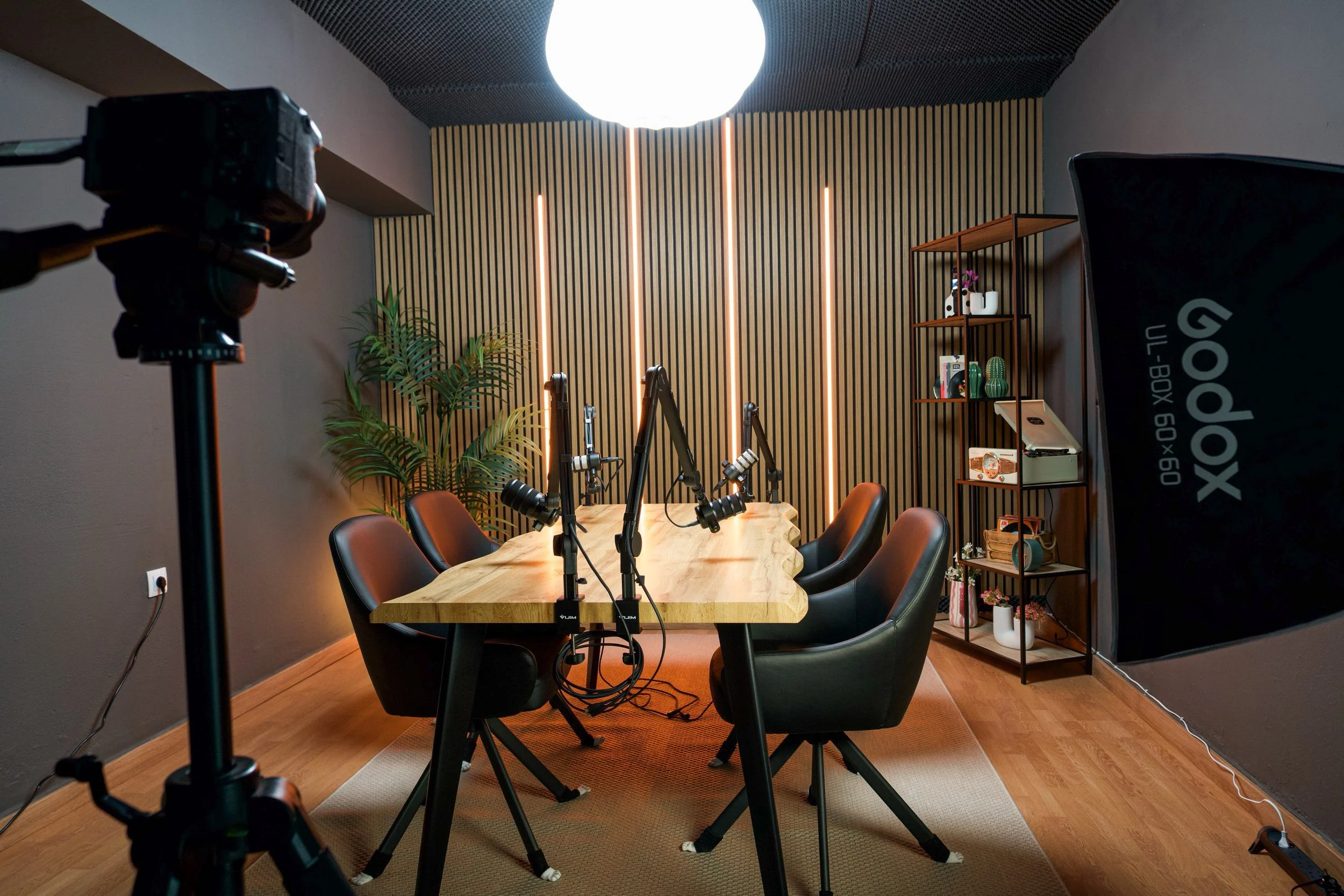 A recording studio or podcast room with a wooden table, six black chairs, a wall with vertical wood slats and LED strips, a plant, a shelving unit with decorative items, and professional microphones set up on the table.