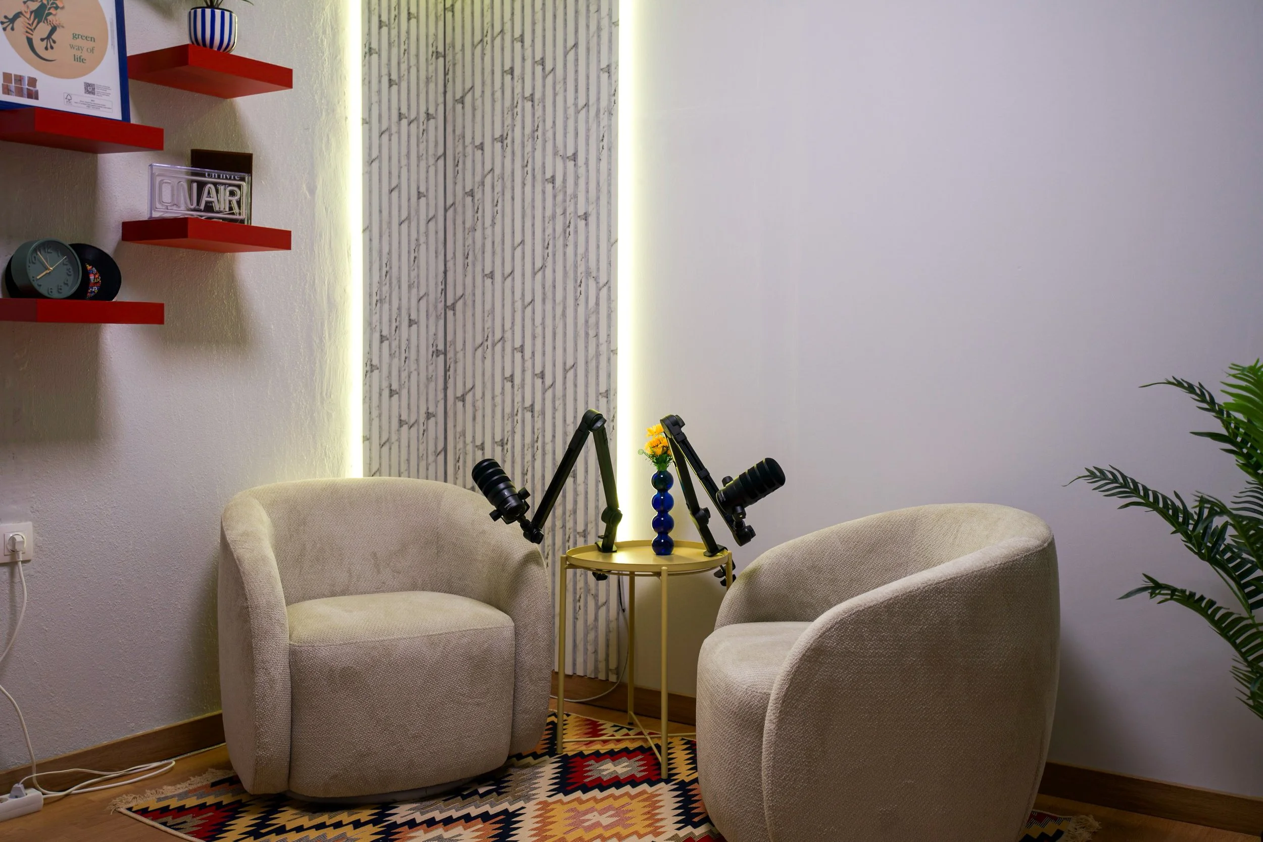 A cozy seating area with two beige armchairs arranged around a small yellow table. The table holds a decorative blue vase with yellow flowers and a black device with adjustable arms. Behind the seating area, there is a wall with a marble strip and vertical lighting, along with a potted plant on the right. On the left, red floating shelves hold decorative items including clocks, a small framed picture, an 