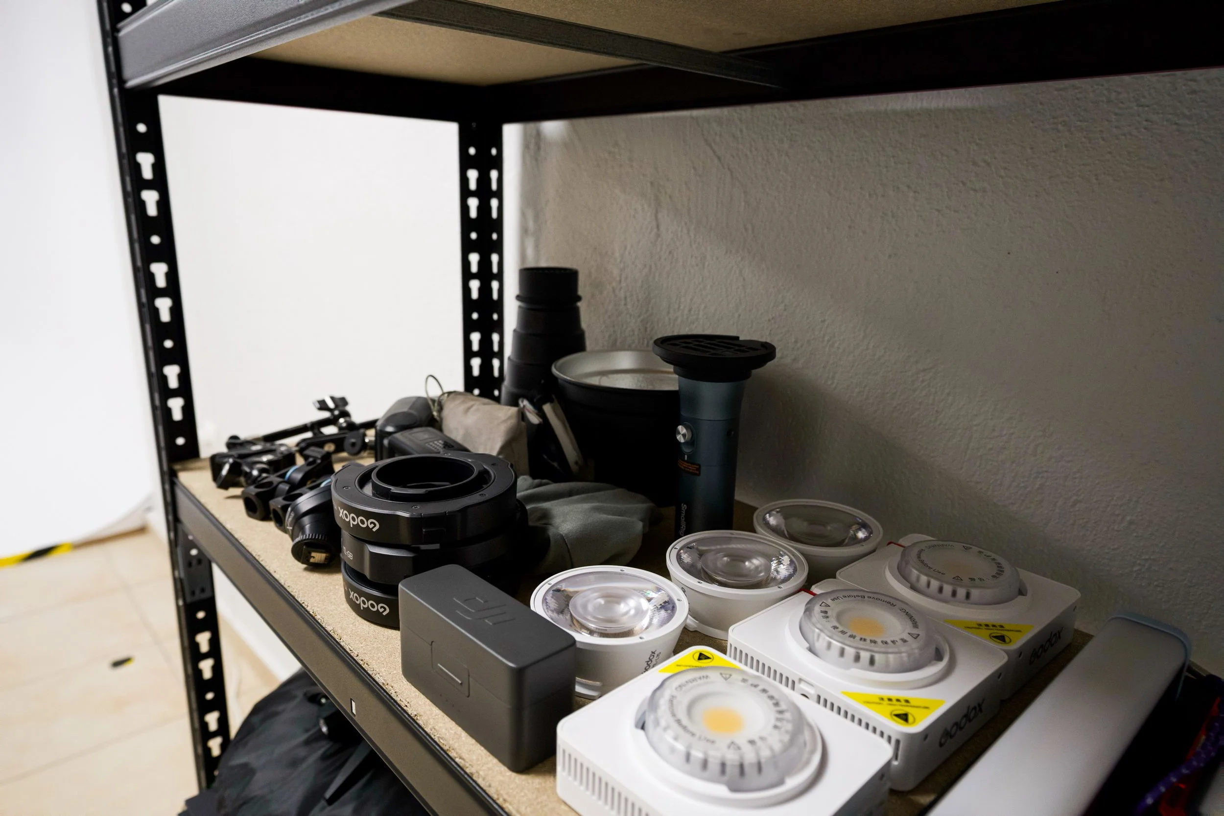 A metal shelf with various equipment and supplies including camera lenses, containers, and filters, set against a plain wall.