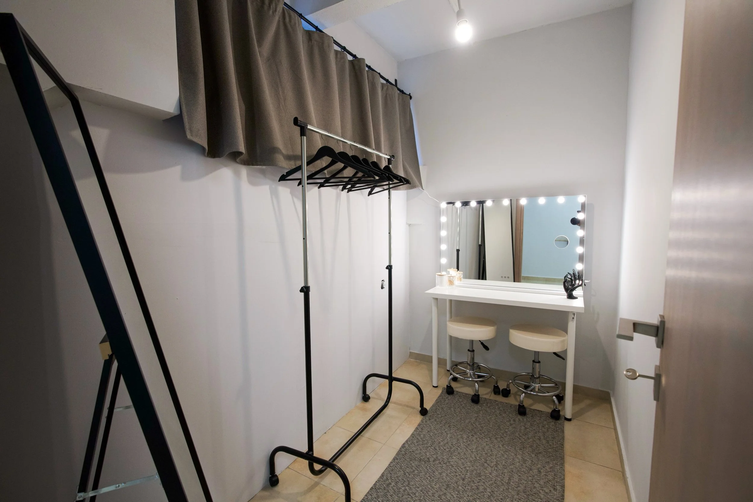 A dressing room with beige walls, a makeup mirror surrounded by lights, a small white table with two beige stools, a clothing rack with black hangers, beige curtains, and a gray floor mat.