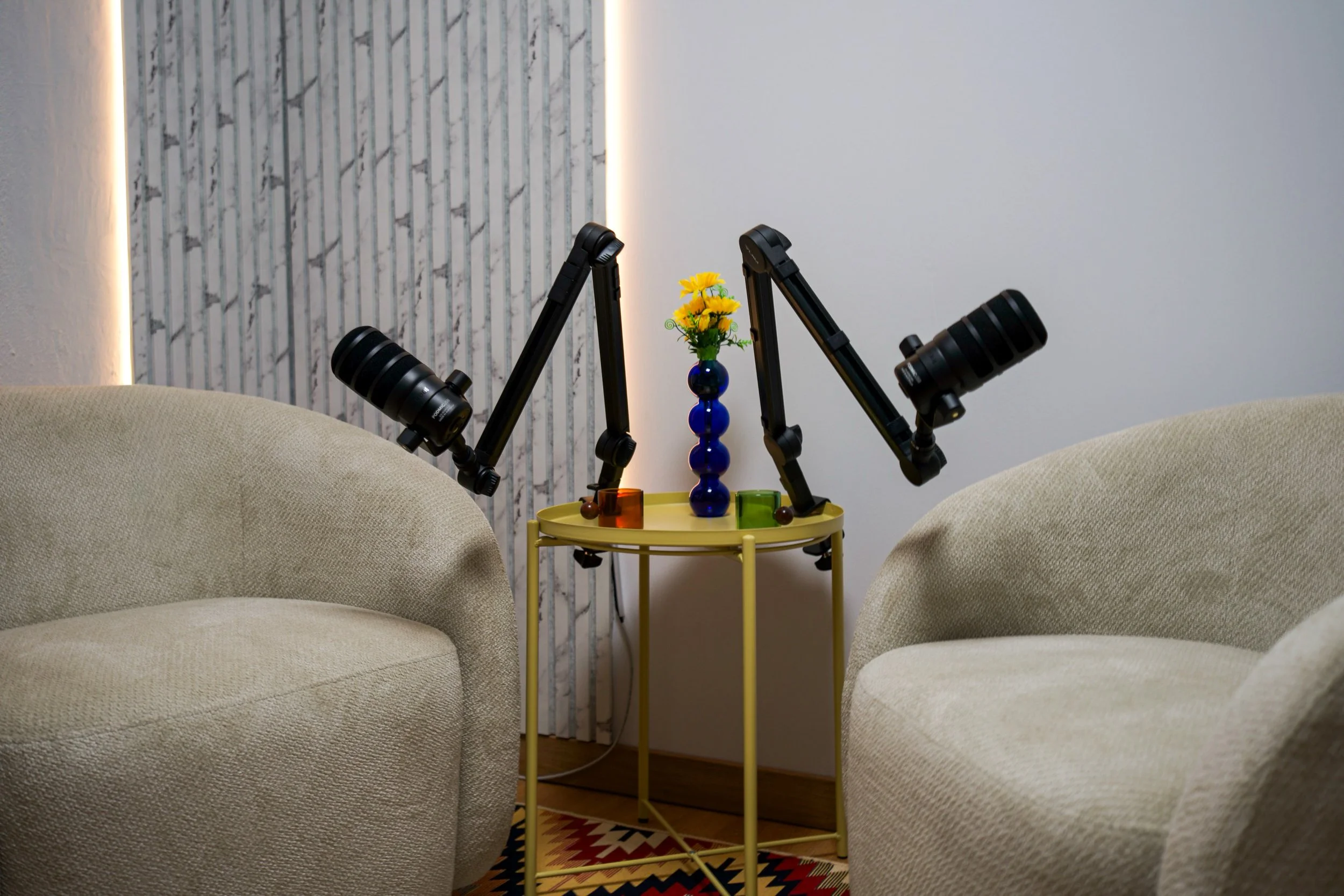 Living room with two beige fabric armchairs facing each other, a yellow round side table between them, with a small blue vase of yellow flowers and colorful decorative objects on it. Behind the table, a wall with vertical marble and wood accents, and a narrow strip of lighting on the left.