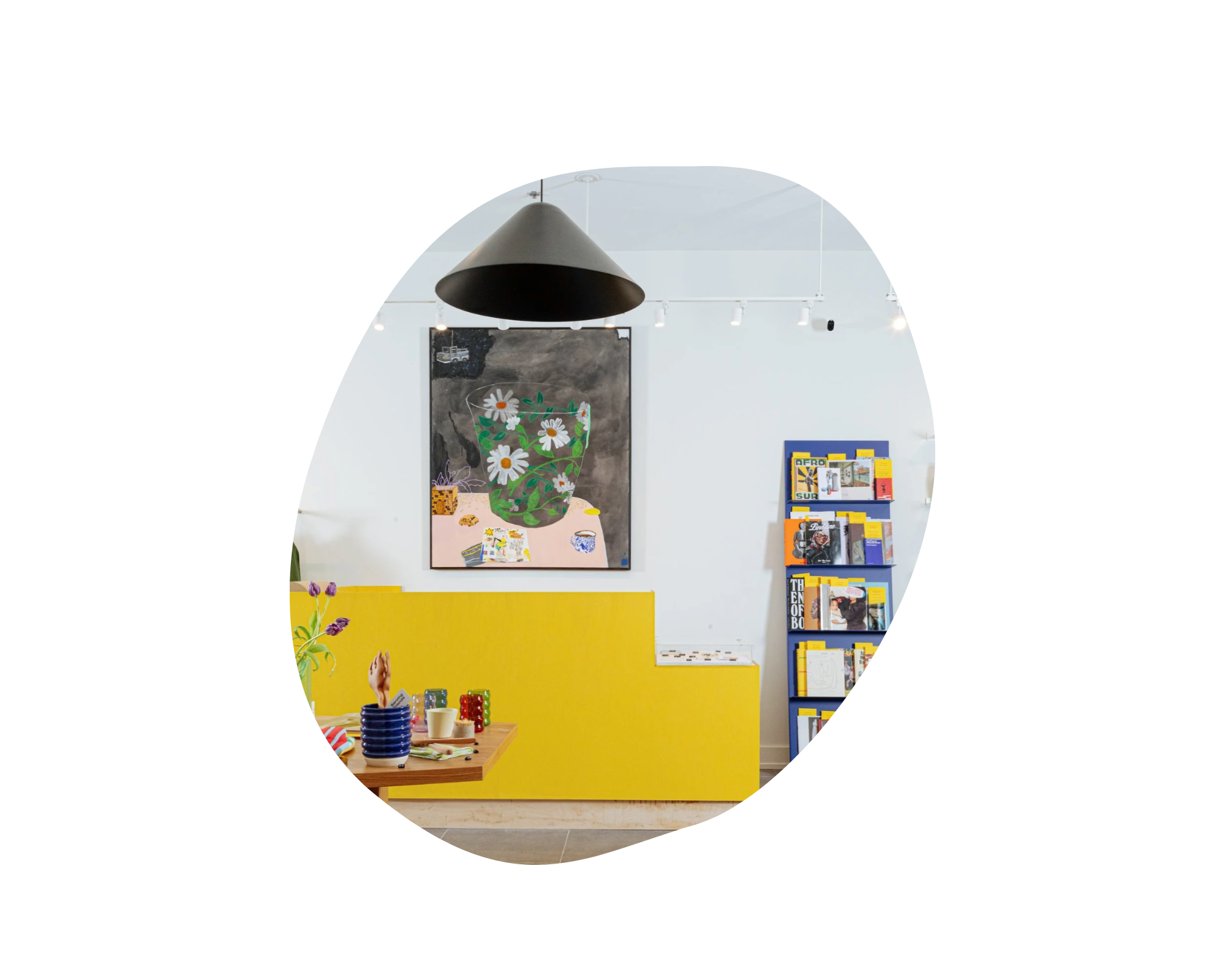 Interior of a modern store or cafe with a black pendant light hanging from the ceiling, a large framed art piece of daisies, a blue bookshelf filled with magazines or books, a yellow divider, and a wooden table with stacks of colorful bowls and cups.
