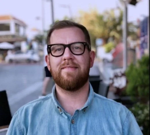 A man with glasses and a beard wearing a light blue button-up shirt outdoors with blurred background of trees, buildings, and boats.