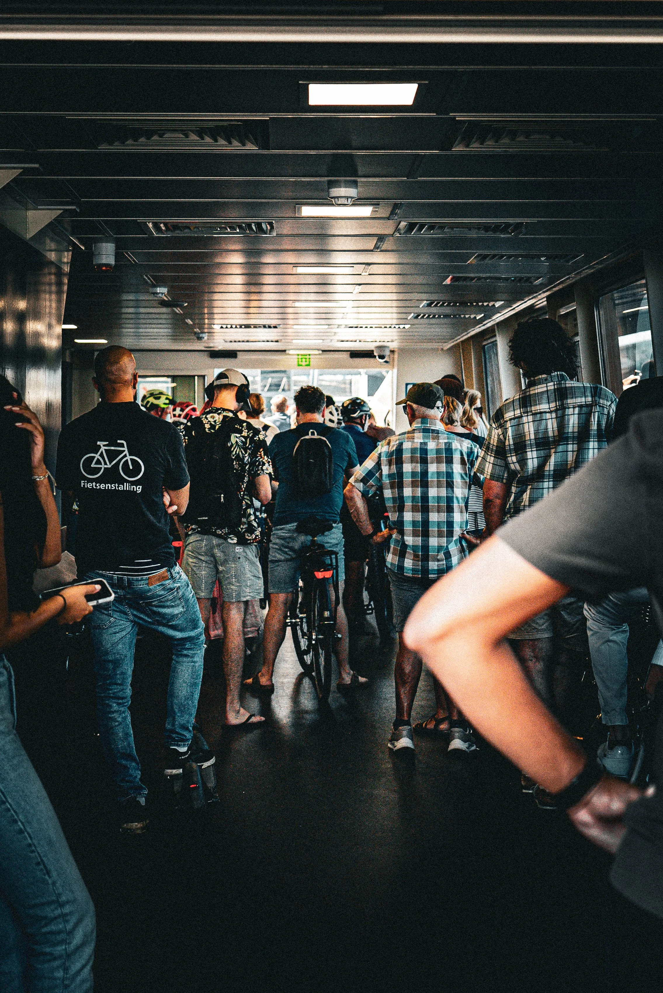 People waiting in a dimly lit corridor with ceiling lights, some with bicycles and backpacks, possibly at a transportation terminal.