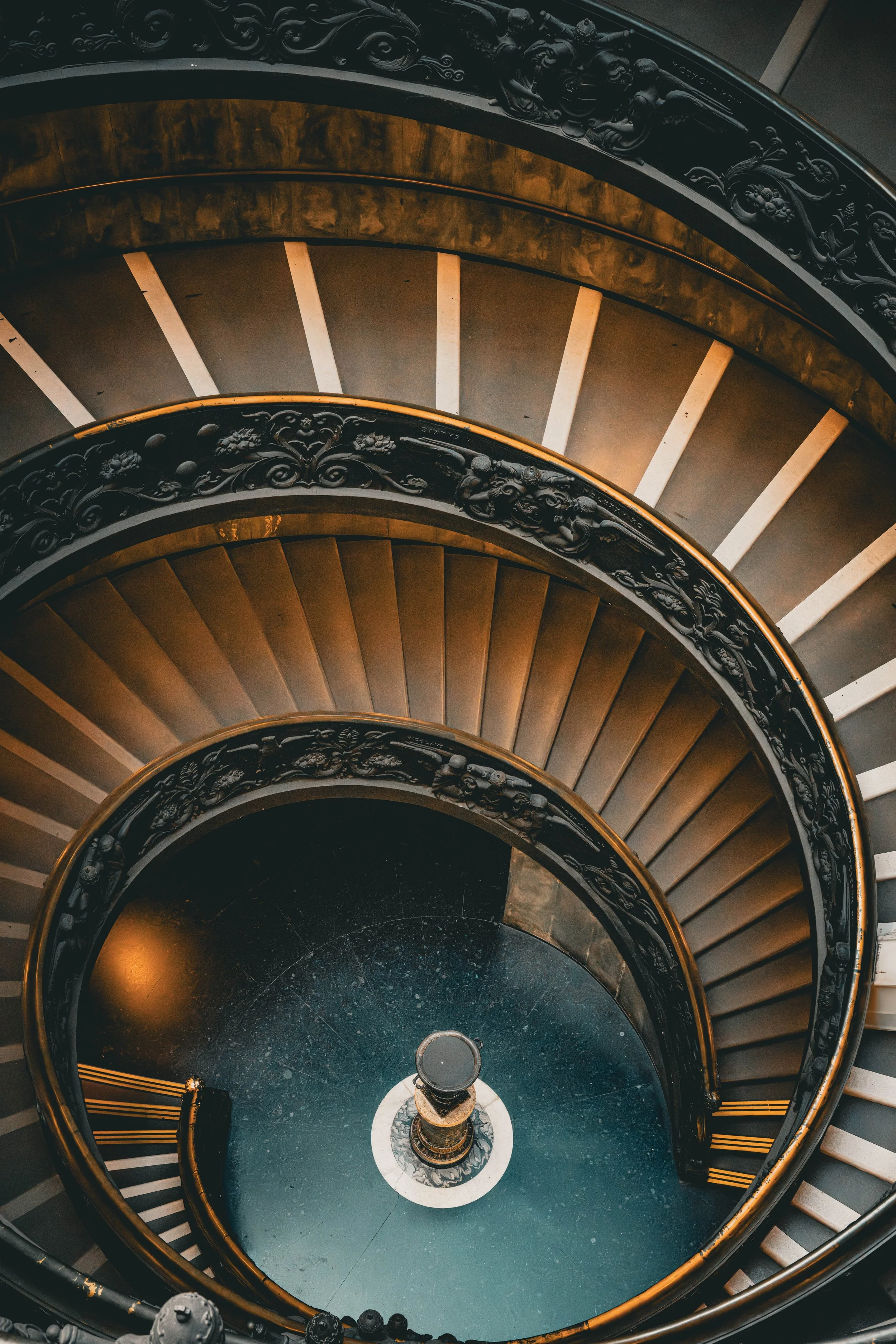 A top-down view of a spiral staircase with ornate black railings and stairs that gradually narrow as they descend to the bottom, where a small circular platform is visible.