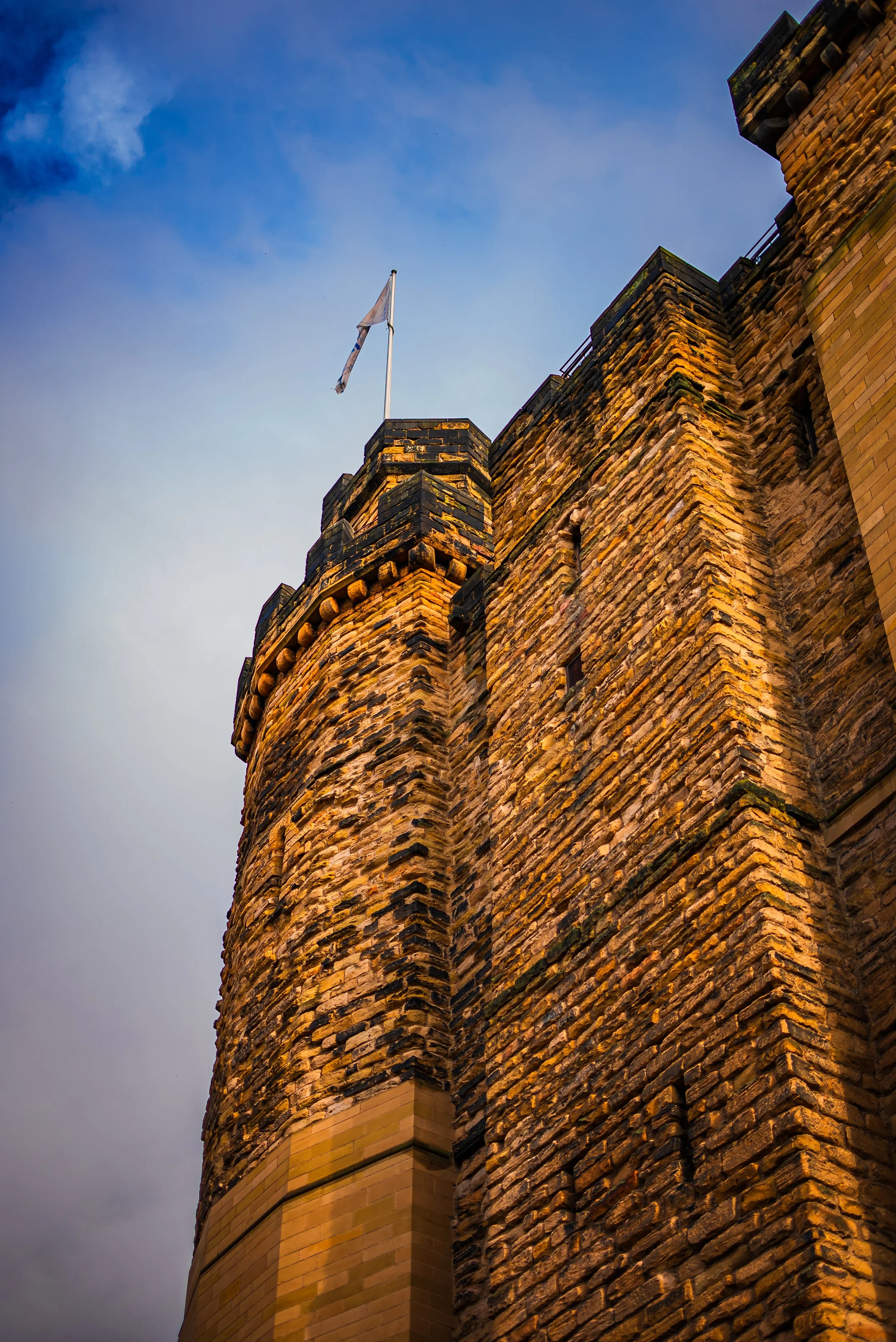 Close-up of a tall, historic stone castle tower with a flag on top, against a partly cloudy sky.
