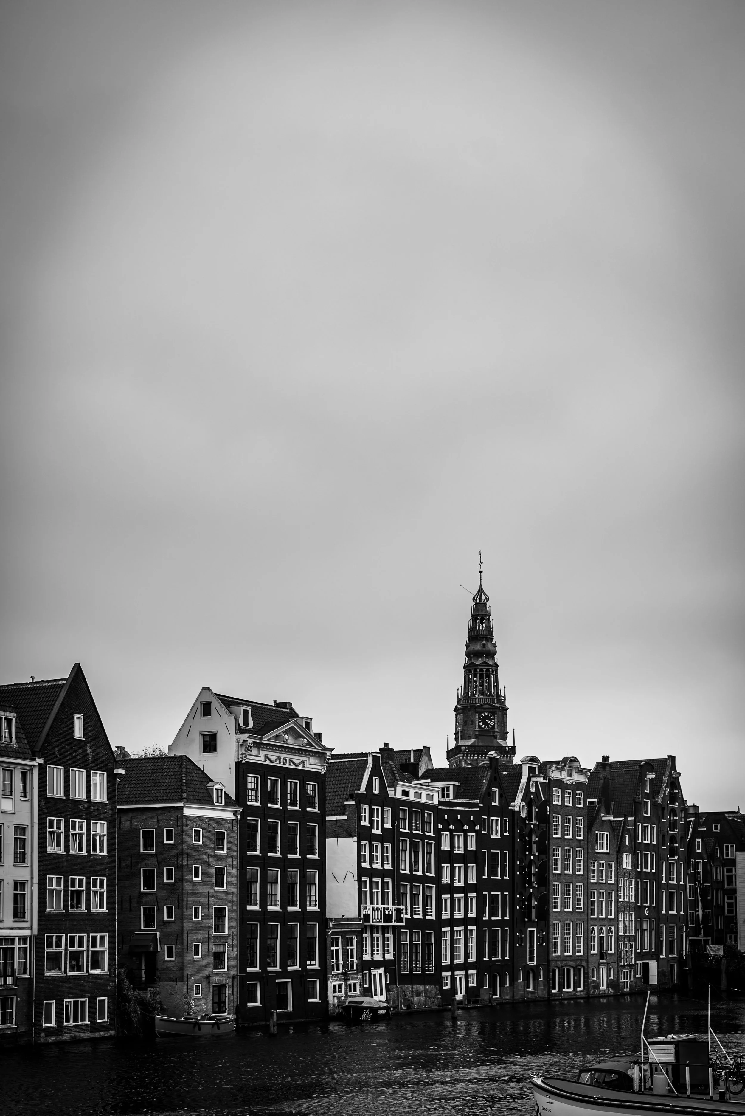 Black and white photo of traditional European buildings along a canal, with a tall clock tower in the background under a cloudy sky.