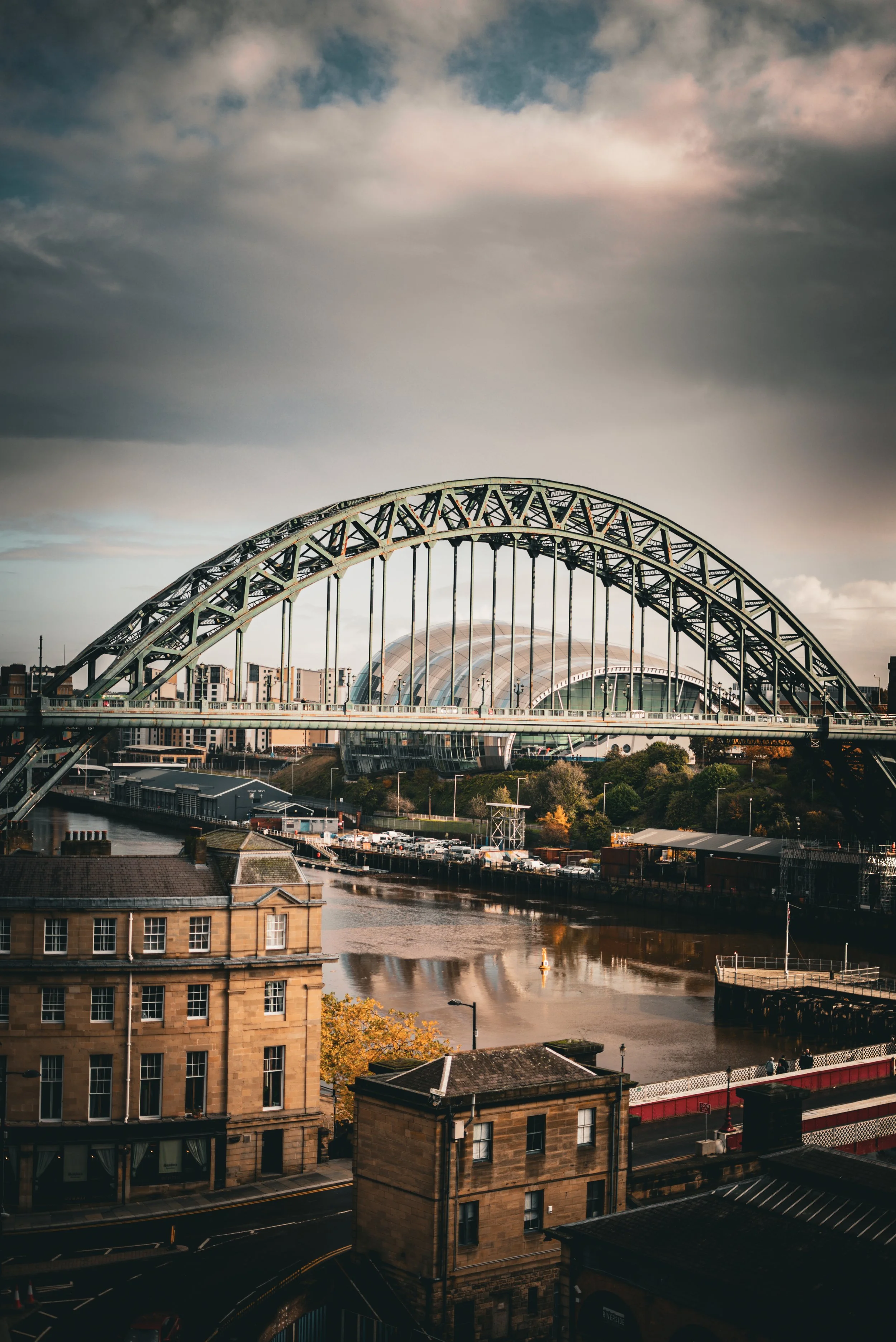 A cityscape with a large arch bridge over a river, with modern architectural buildings and cloudy sky in the background.