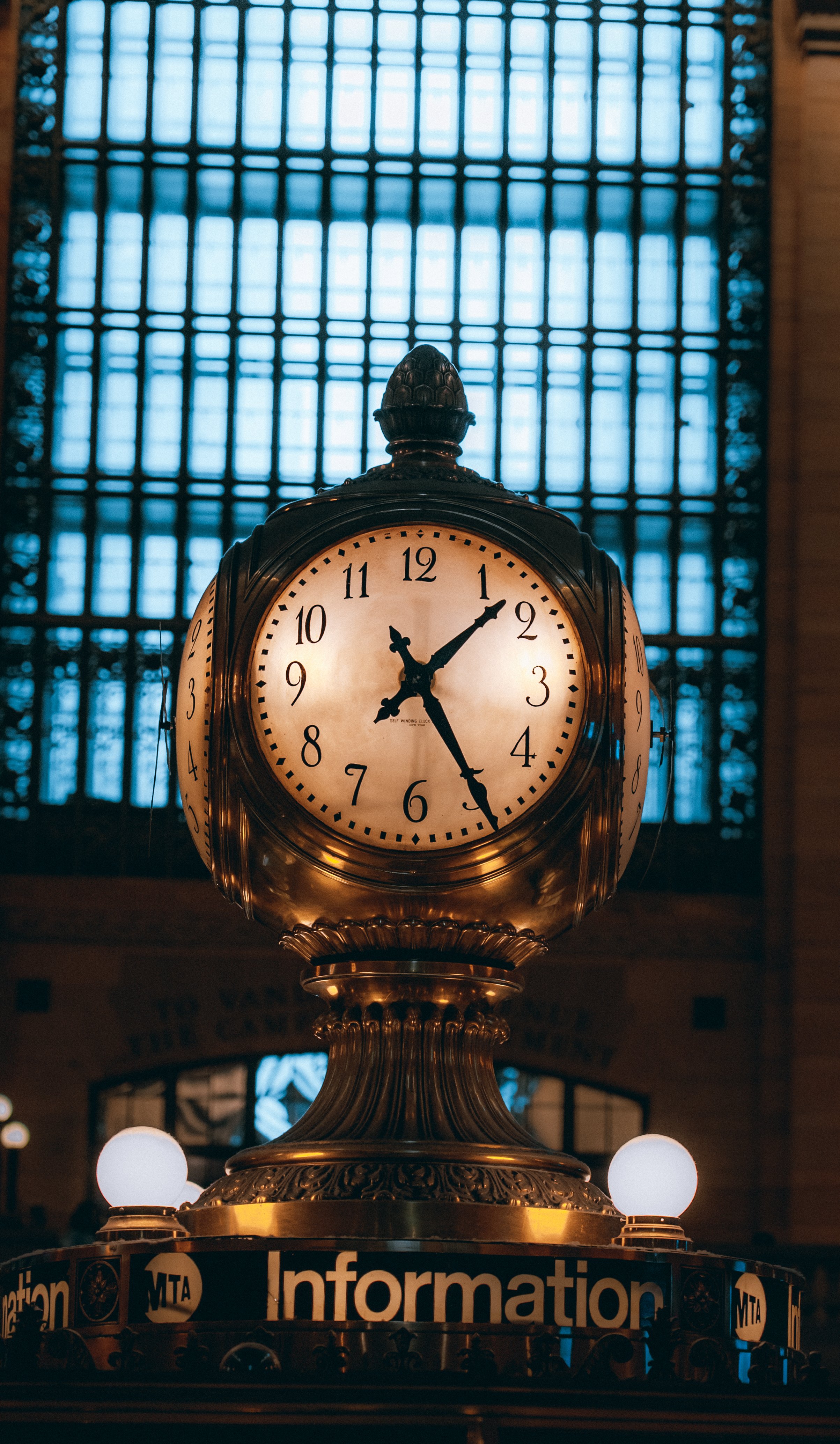 A vintage clock showing 1:24, situated in a train station with a large window behind and a wooden interior.