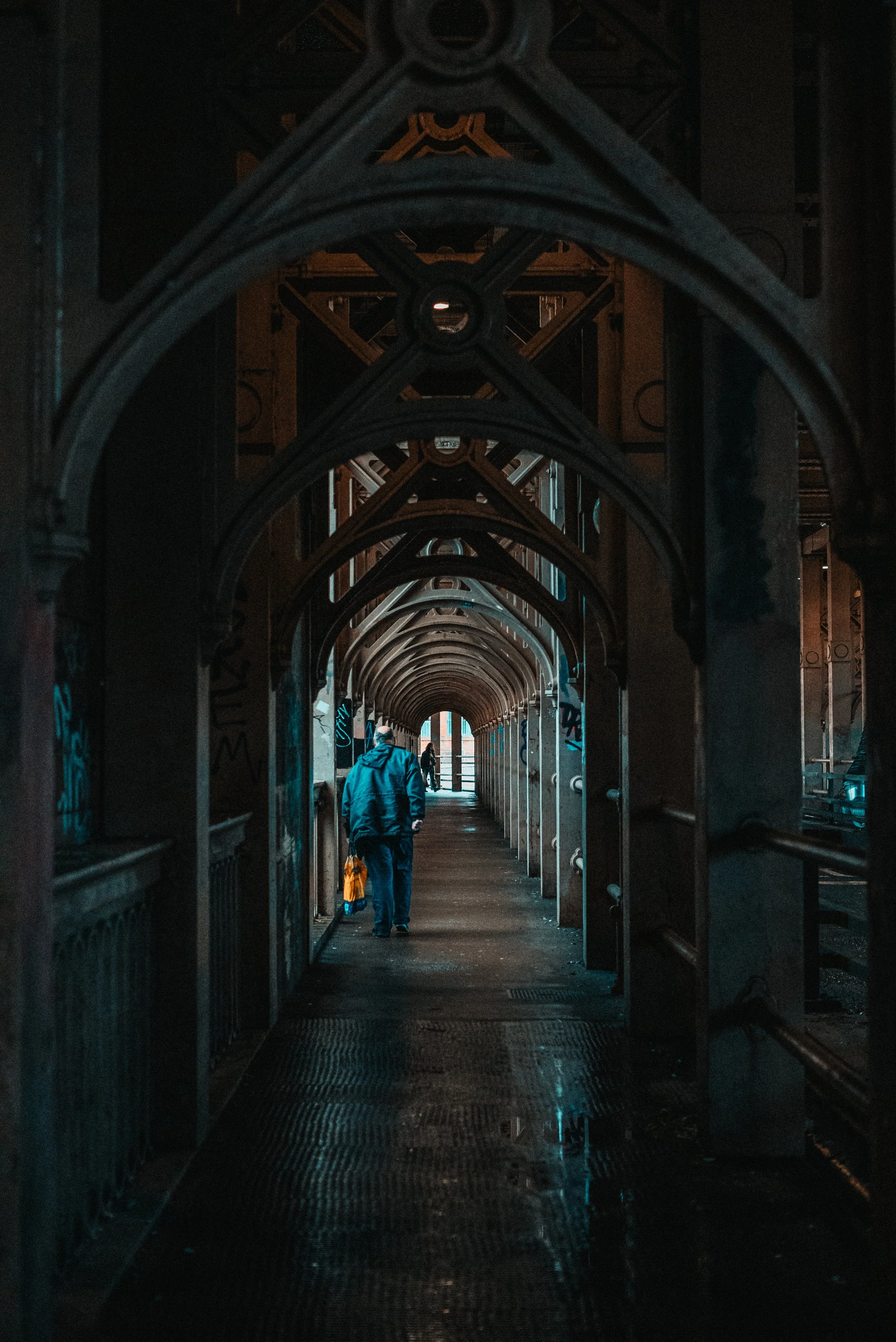 A person walking under an arched, Gothic-style metal walkway in an urban setting, with graffiti on the walls and subdued lighting.