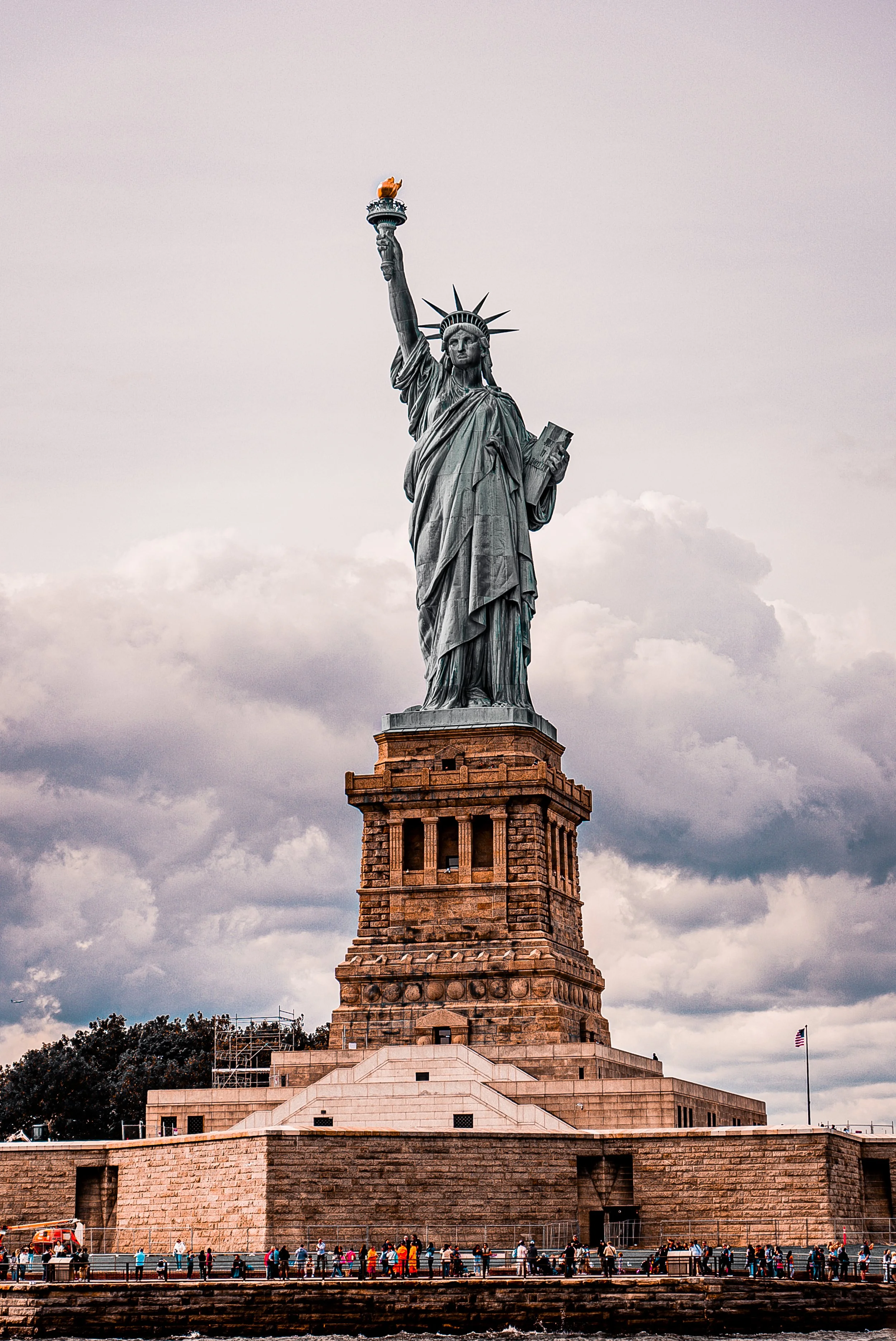 The Statue of Liberty standing tall on Ellis Island with its torch raised and a tablet in its other hand under a cloudy sky.