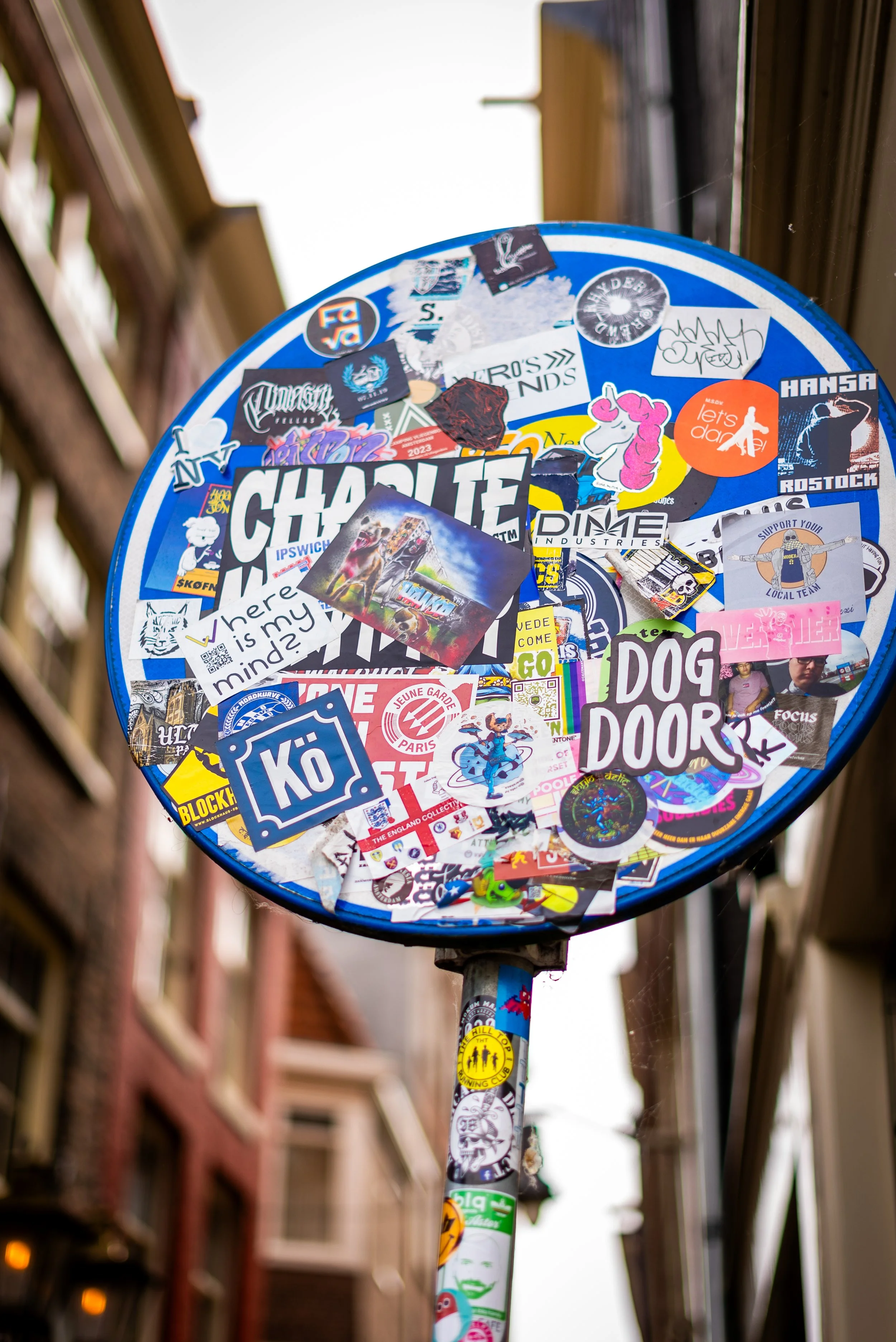 Close-up of a blue pole with a circular sign covered in various stickers and patches, including text, logos, and images, with a blurred city street background.