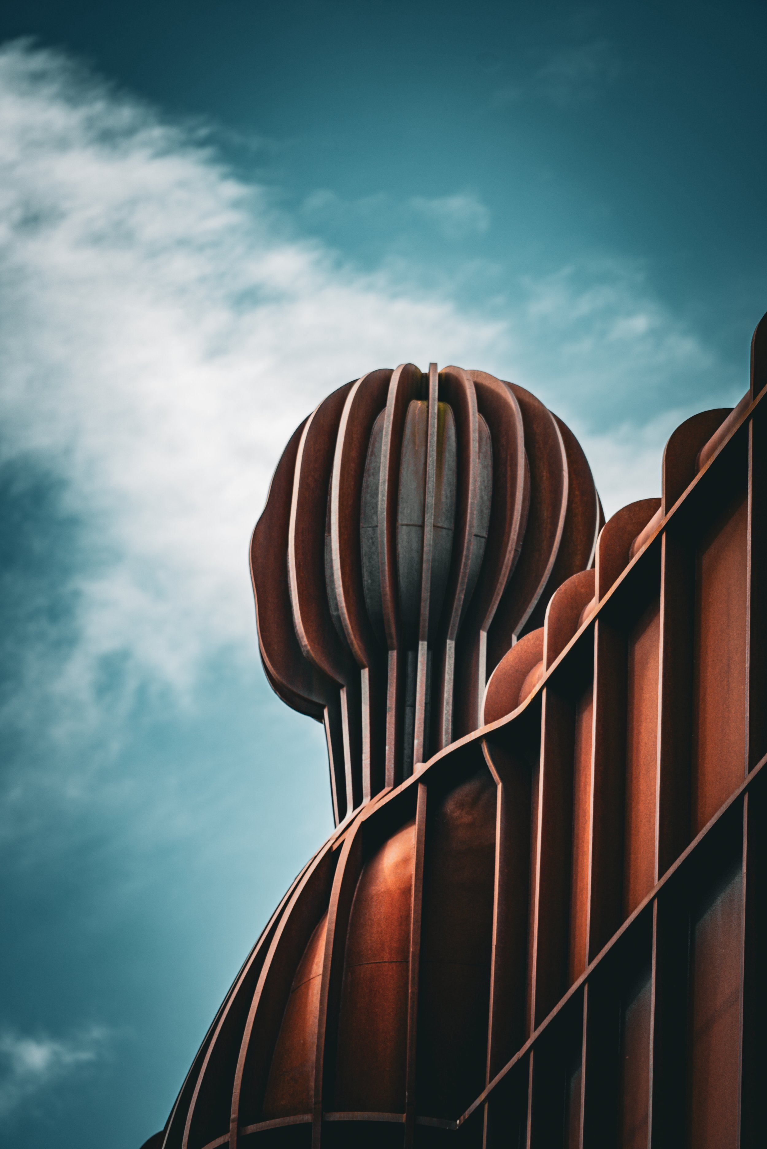 Close-up view of a modern architectural building with a rounded, layered, rust-colored exterior against a partly cloudy sky.