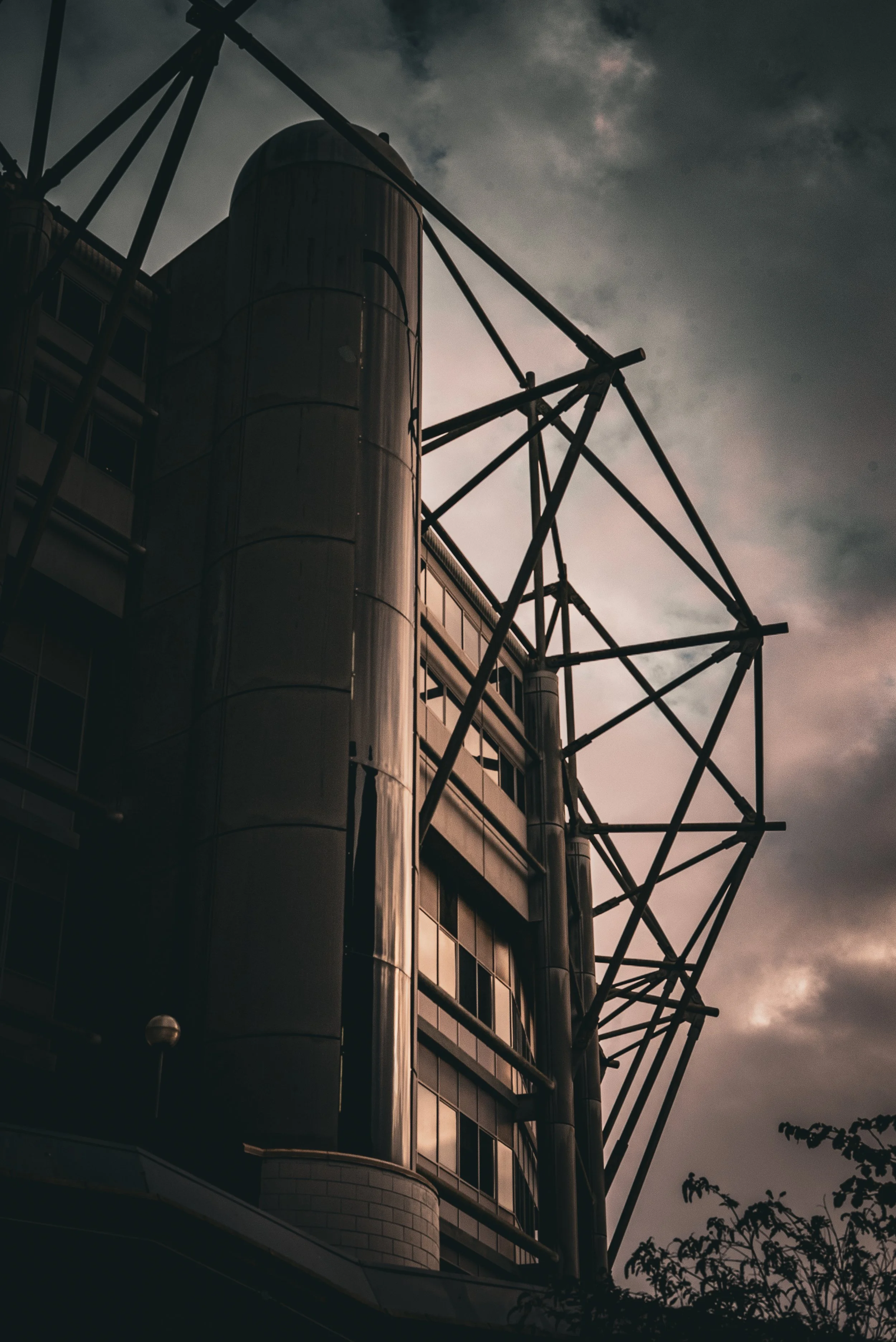 A modern building with metallic surfaces and large windows, featuring a steel framework extending from the top, set against a cloudy sky at dusk.
