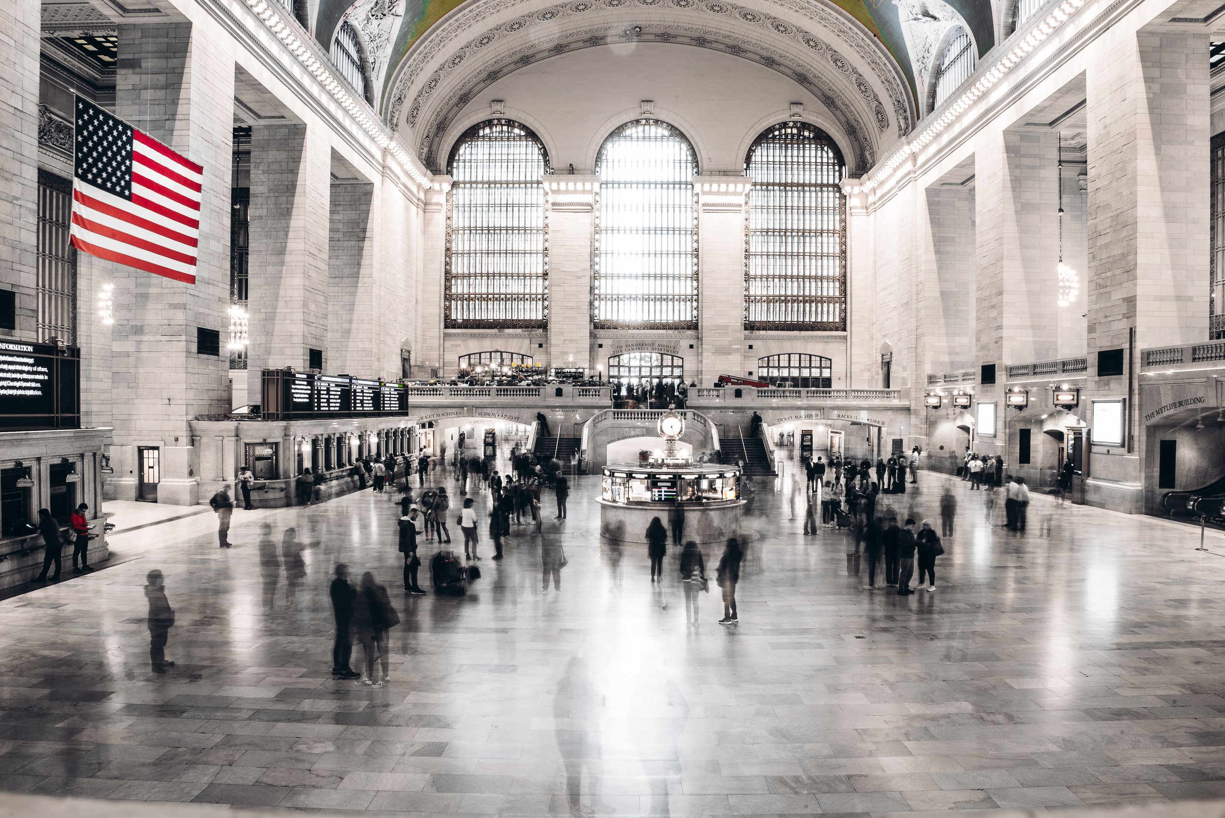 Interior of a grand train station with high arched windows, a large American flag hanging on the left, and people walking or standing around the spacious marble floor.