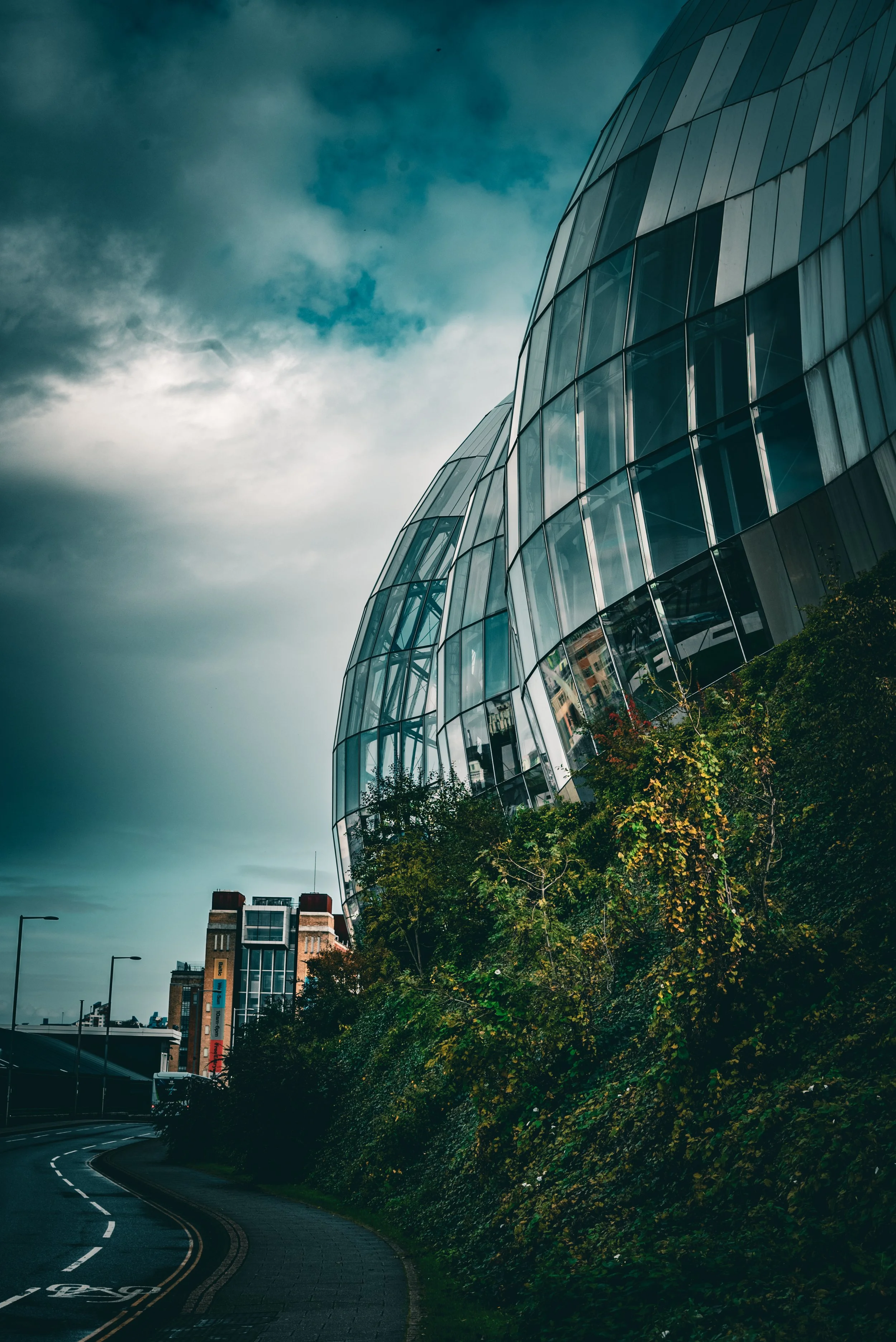Modern glass building with a curved facade reflecting the cloudy sky and nearby structures, alongside a winding road with greenery on the side.