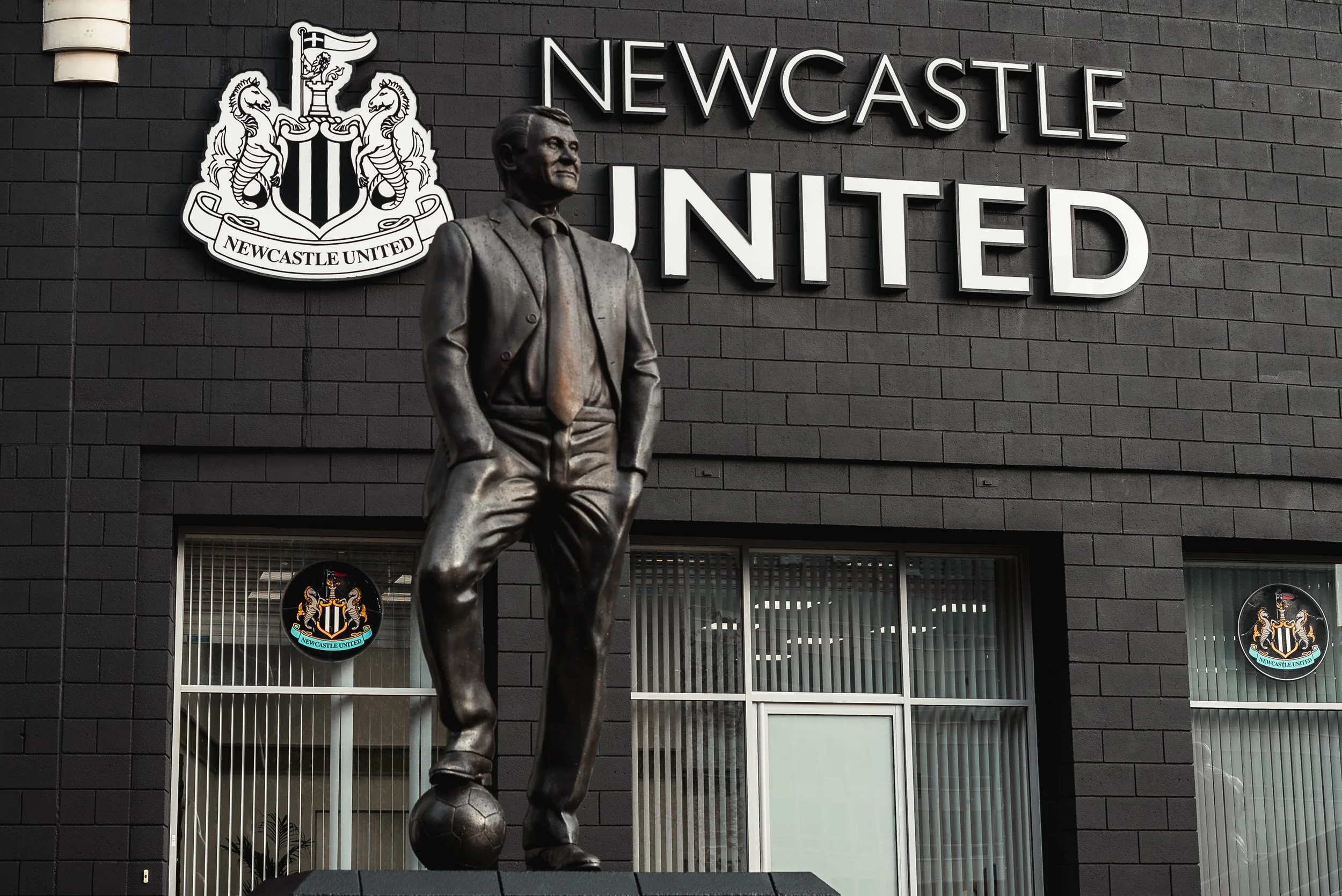 Statue of a man in a suit standing with one foot on a soccer ball in front of a Newcastle United building, featuring the club's emblem and large signs that read "Newcastle United."