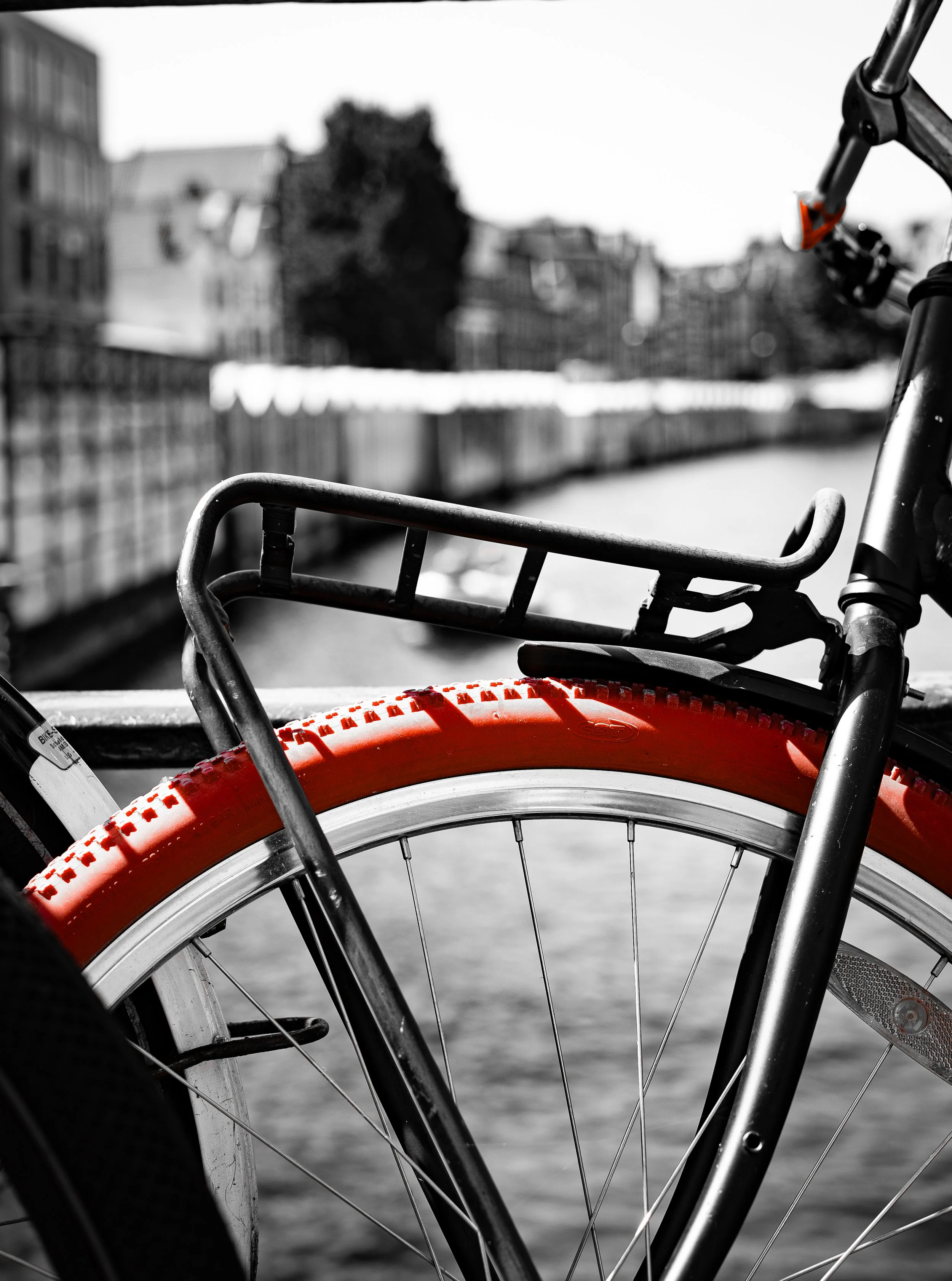 Close-up of a bicycle with a red tire and metal frame, set against a blurred background of a river and buildings.