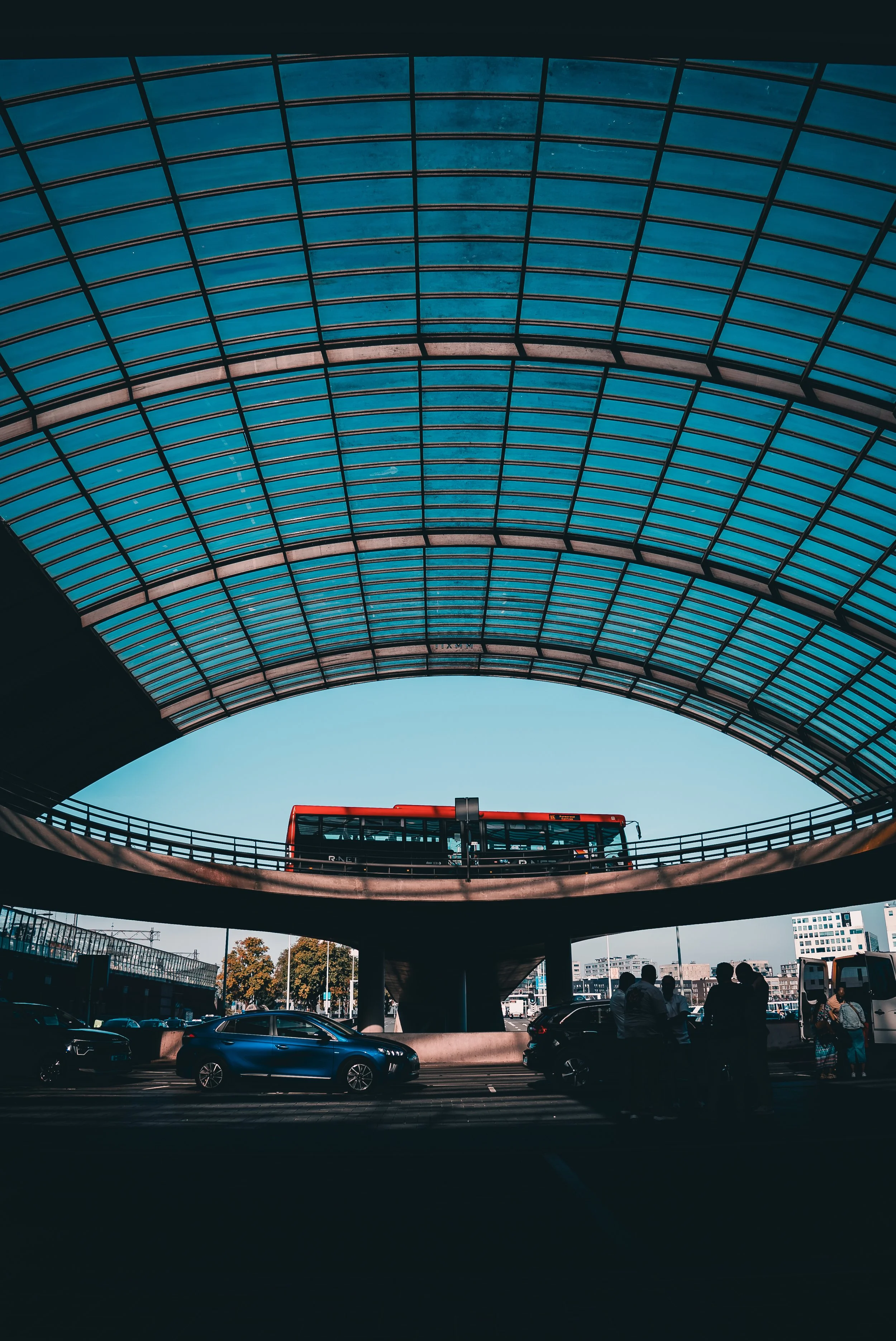 A modern bus station with a curved glass roof and an elevated bus traveling on a circular track, with parked cars and people underneath.