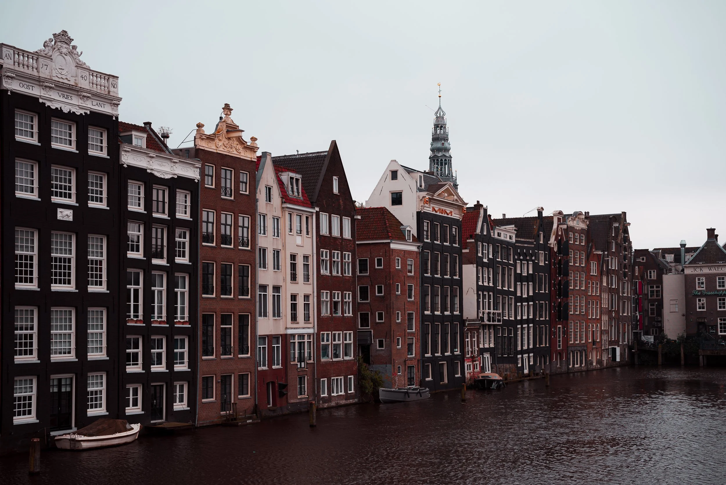 Row of tall narrow European-style buildings facing a canal, with a church steeple in the background, on a cloudy day.