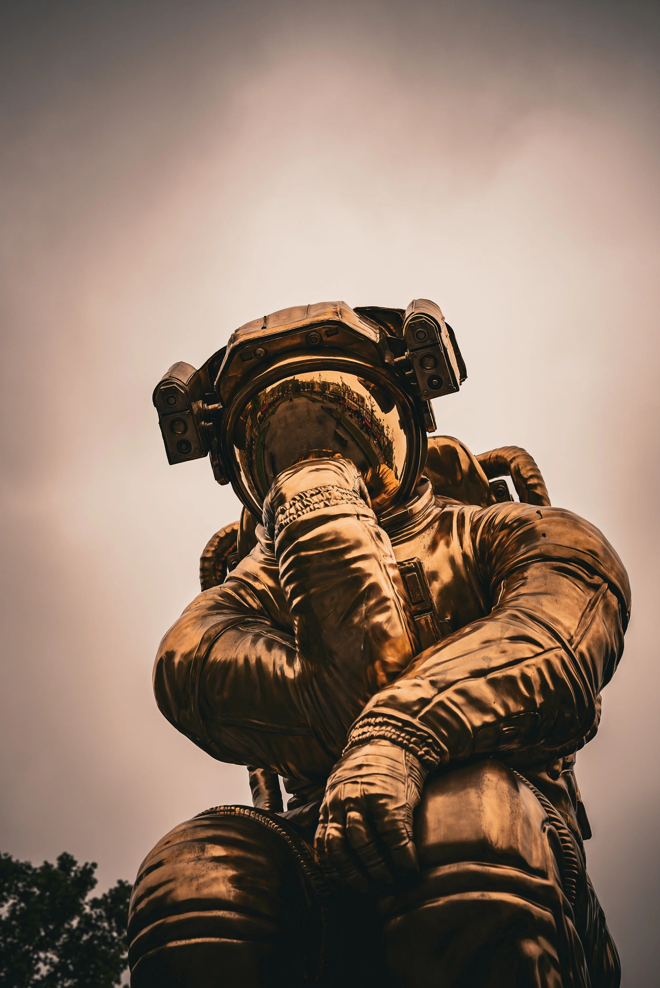 A bronze statue of an astronaut in a reflective helmet, posed with one hand resting on their chin, outdoors with trees and cloudy sky in the background.