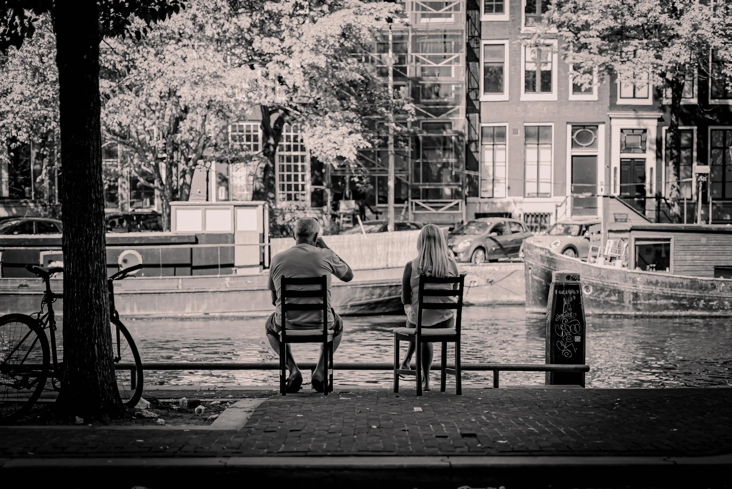Two people sitting on chairs by the water, facing away. One is an older man and the other is a woman. There is a bicycle on the left and a boat on the right, with buildings and trees in the background.