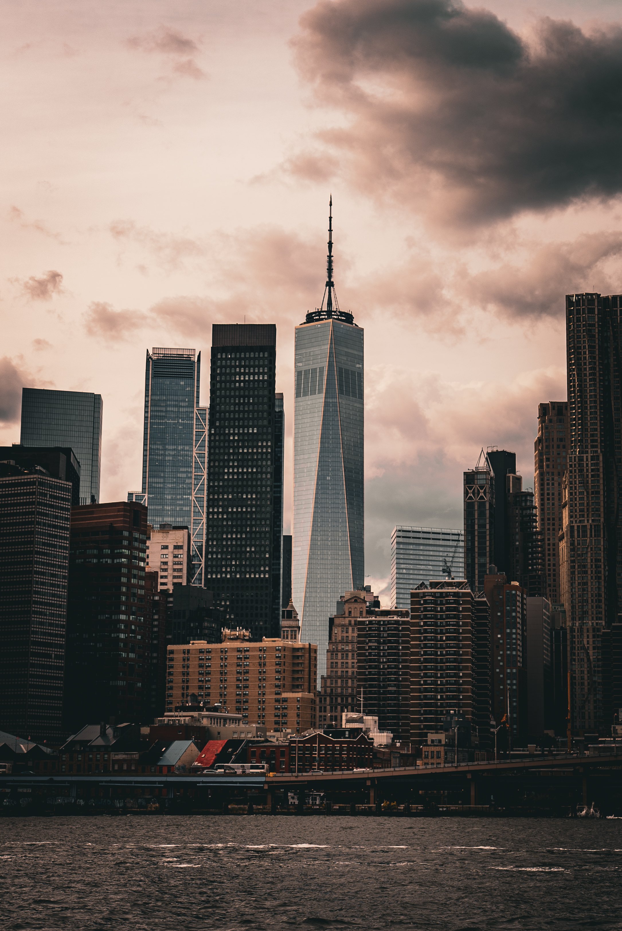 New York City skyline at sunset with One World Trade Center towering among other skyscrapers over the water.