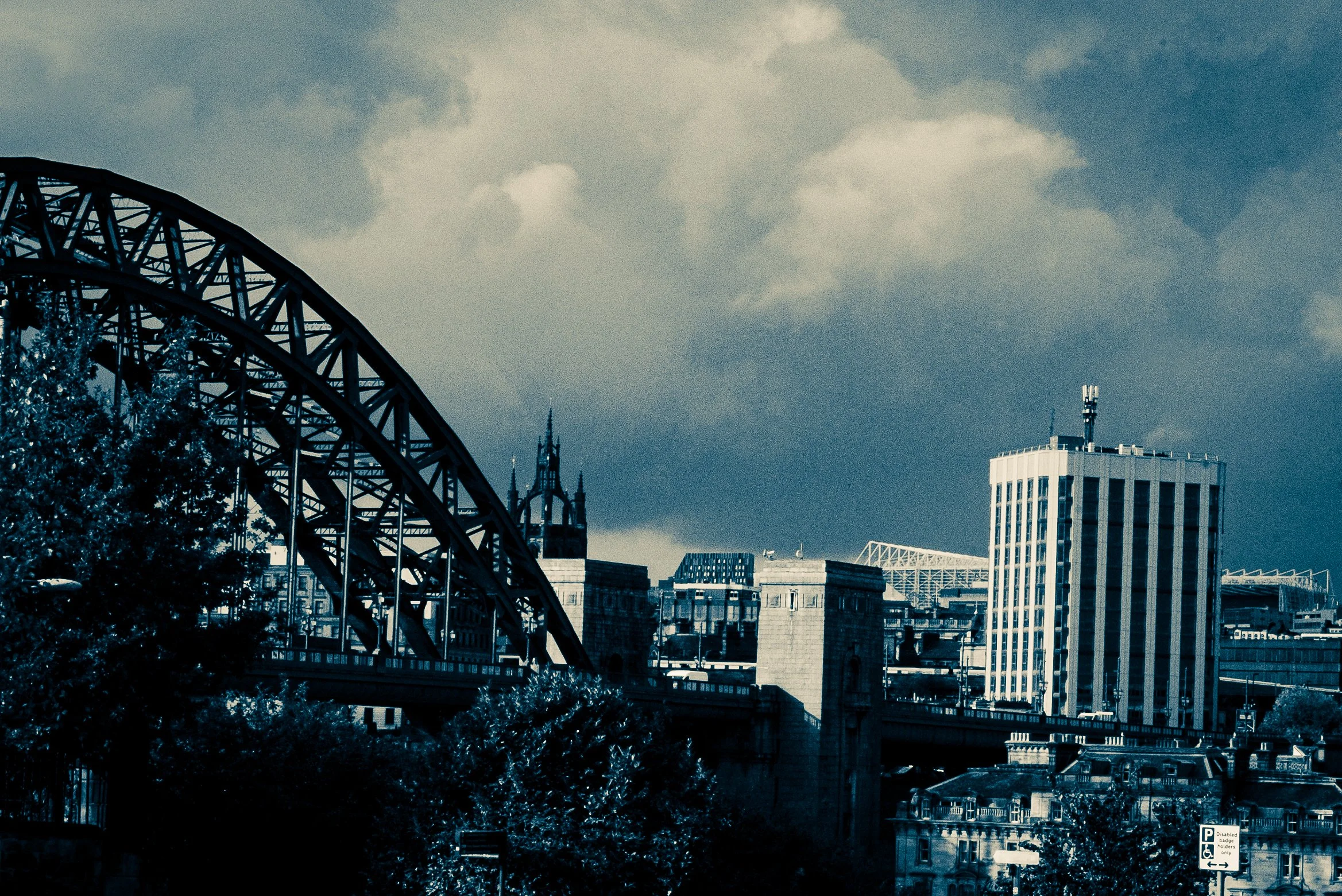 A city skyline with a bridge in the foreground, tall buildings, and a cloudy sky.