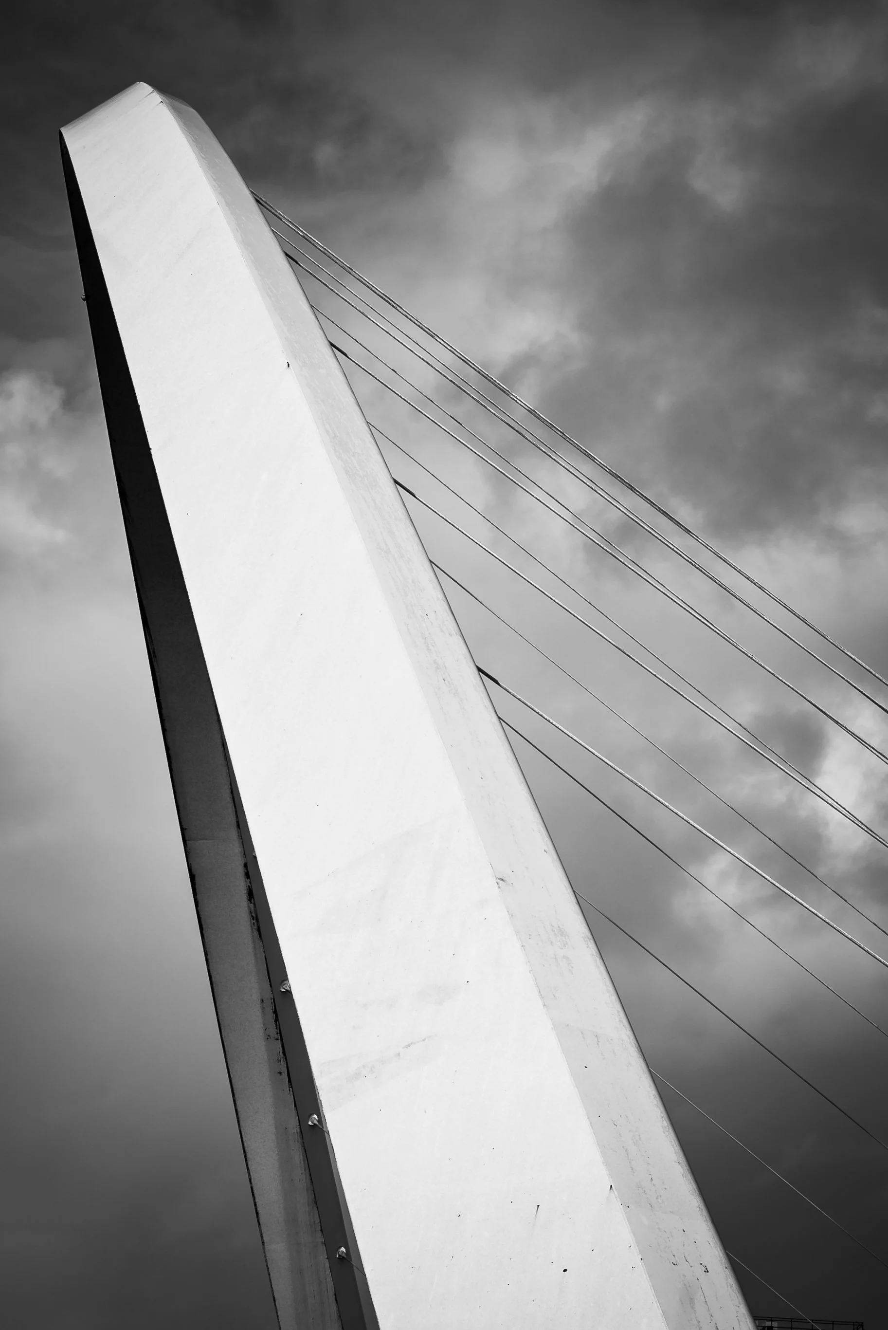 Black and white photo of a tall bridge support with cables extending from it, against a cloudy sky.