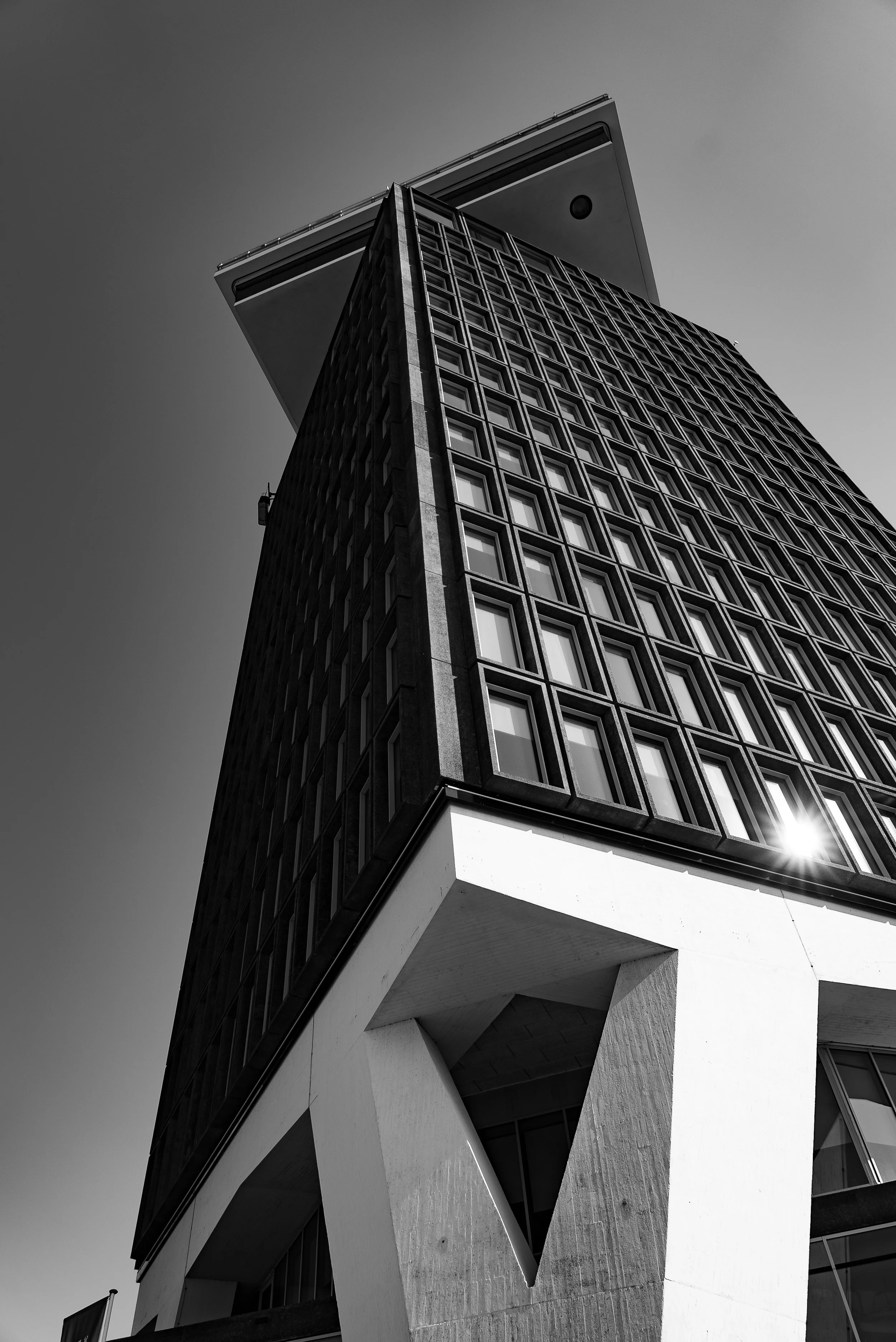 Black and white photo of a modern multi-story building with large windows, viewed from below, with the sunny sky in the background.