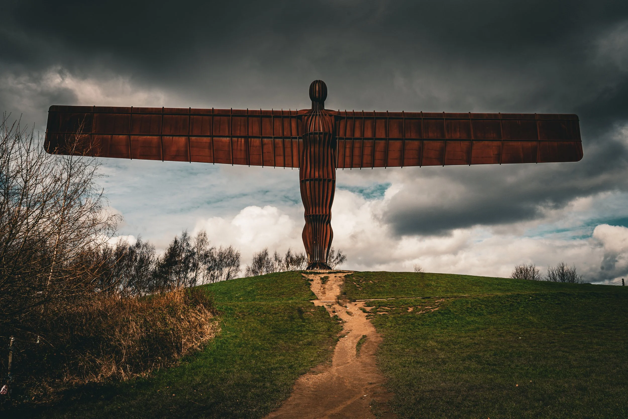 A large sculpture of a human figure with outstretched arms, made of wood or metal, standing on a grassy hill against a dark, cloudy sky.