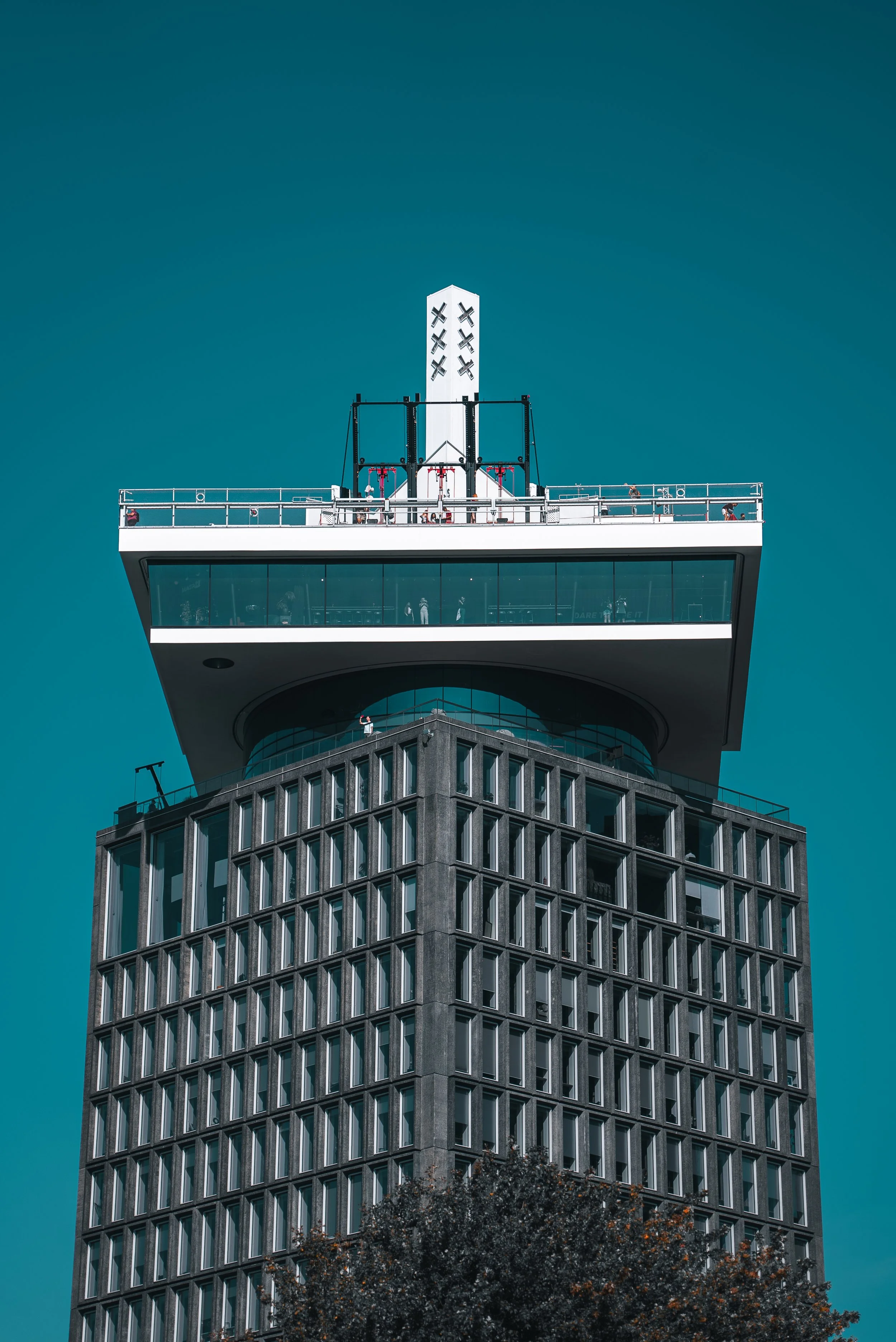 Close-up of a tall modern building with a distinctive overhanging structure at the top, featuring large windows and a dark gray exterior, against a clear bright blue sky.