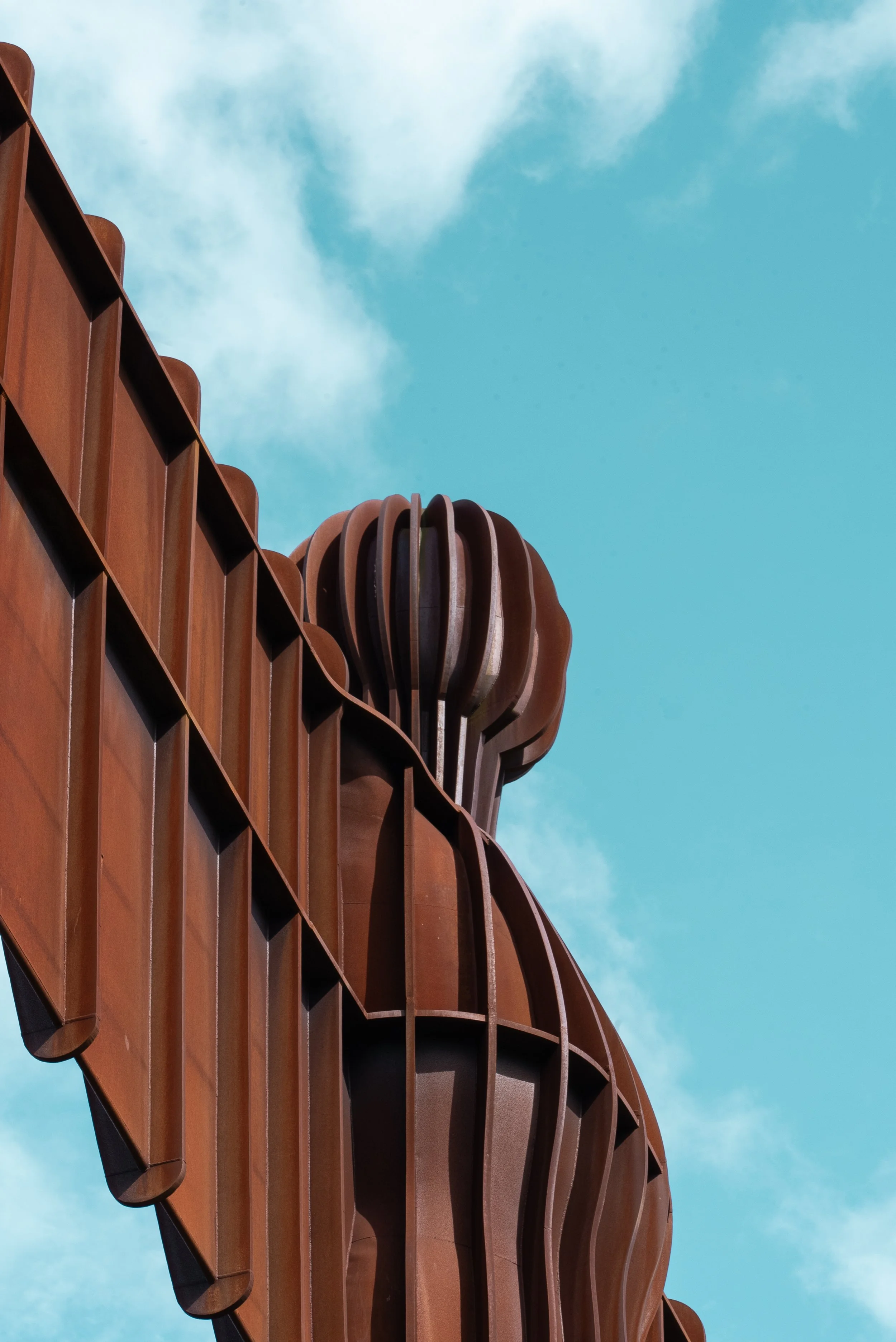 Close-up of a modern architectural structure made of rust-colored metal, featuring curved and vertical elements, against a blue sky with some clouds.