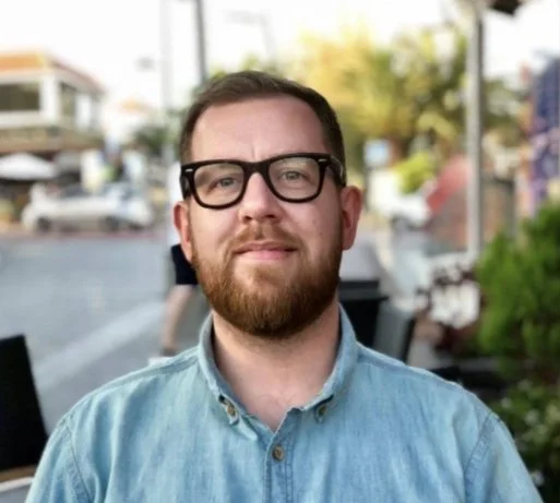 A young man with glasses and a beard, wearing a light denim shirt, standing outdoors on a city street with blurred background.