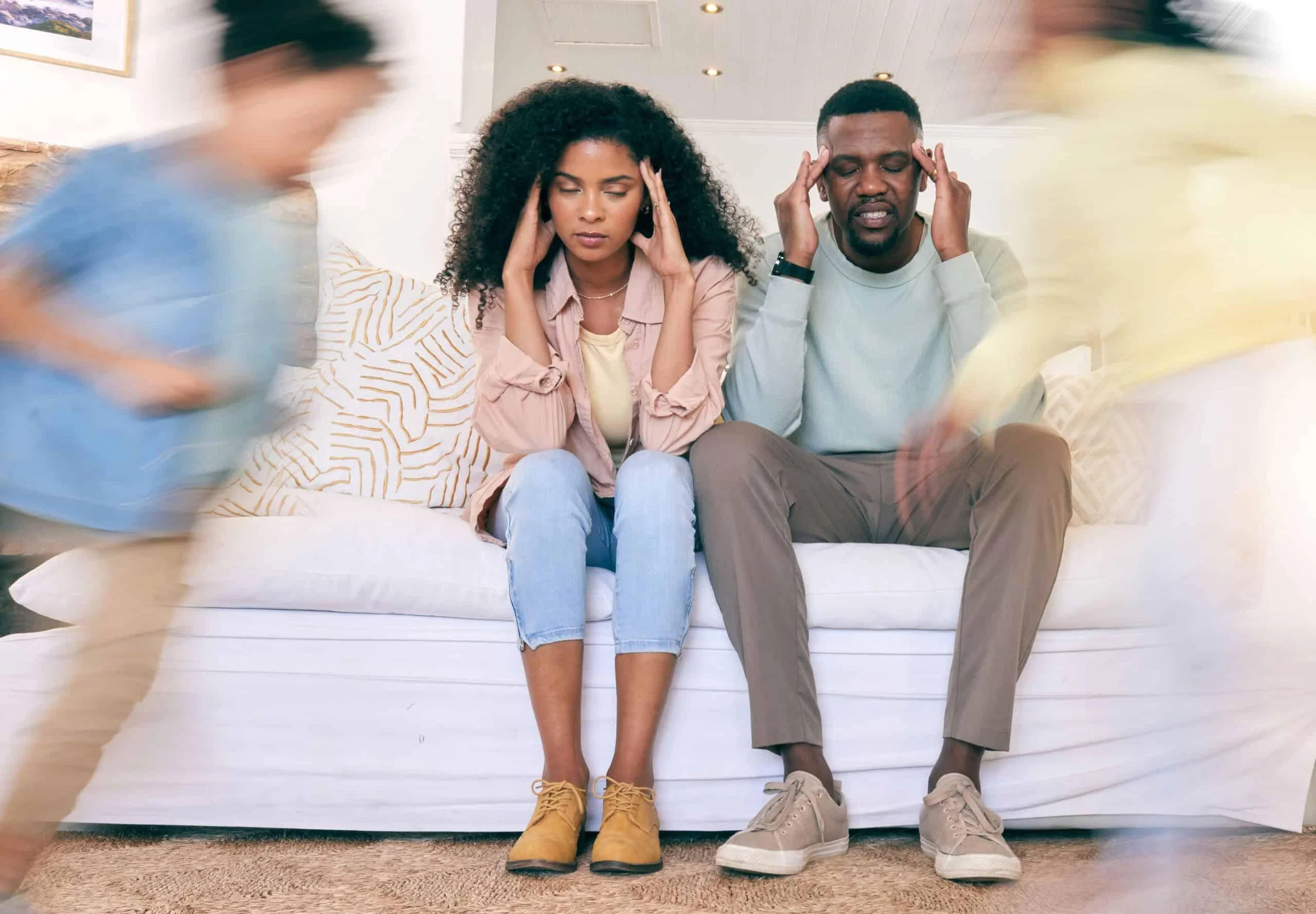 A woman and a man sitting on a couch with a distressed look, holding their heads in their hands. Two blurred figures walk past in front of them.